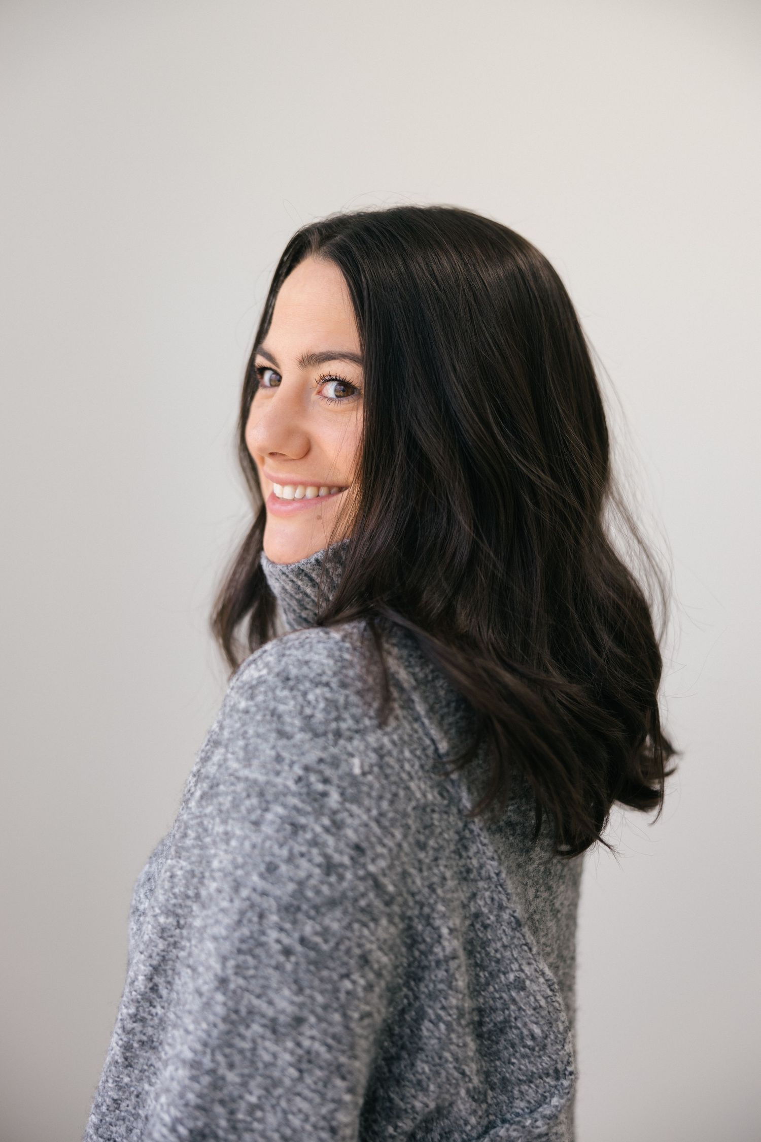 Woman with dark wavy hair wearing a cozy gray turtleneck sweater poses against light background in photo series.
