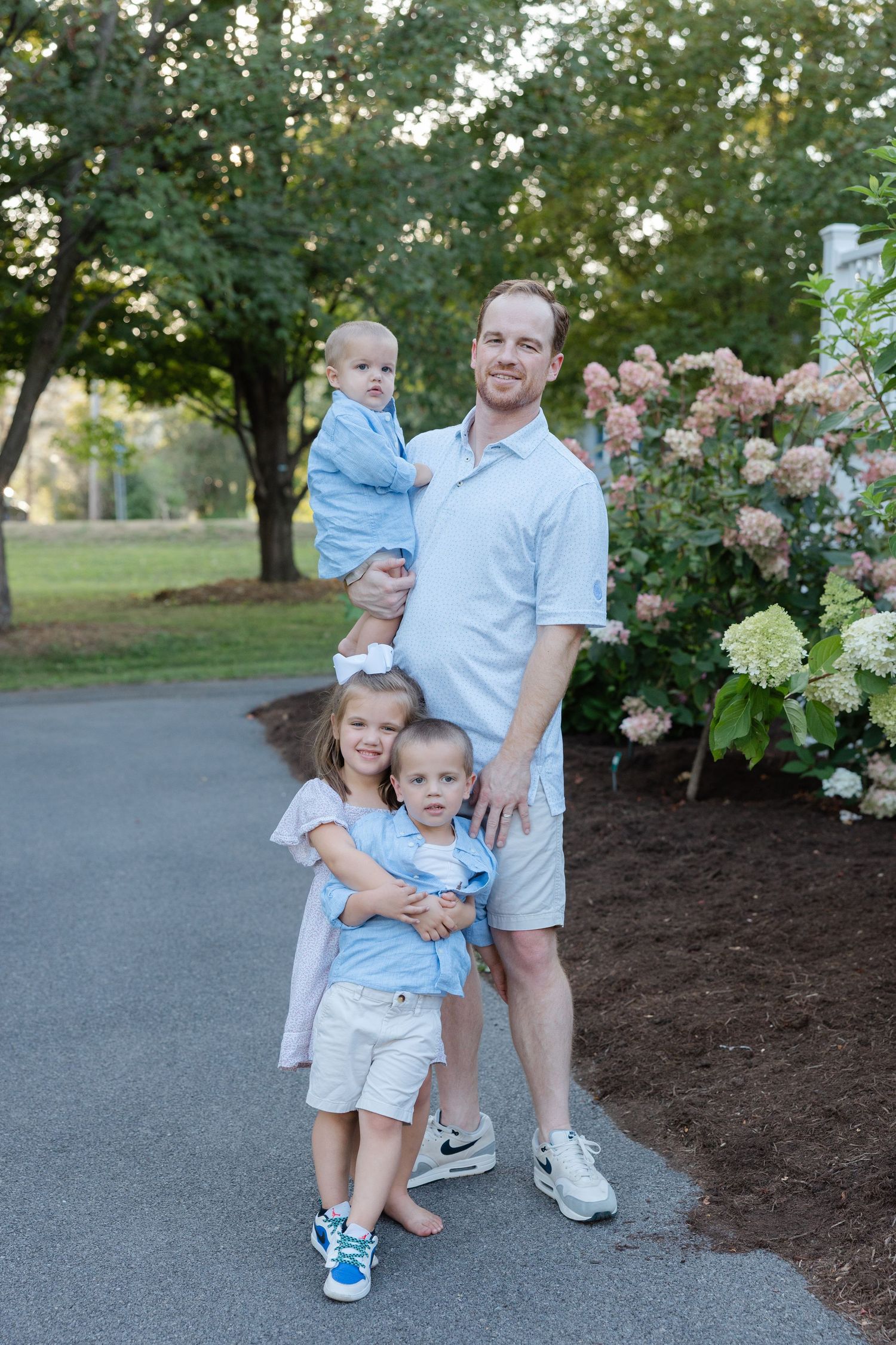 A family poses together on a paved path beside blooming hydrangea bushes and trees in a garden setting.
