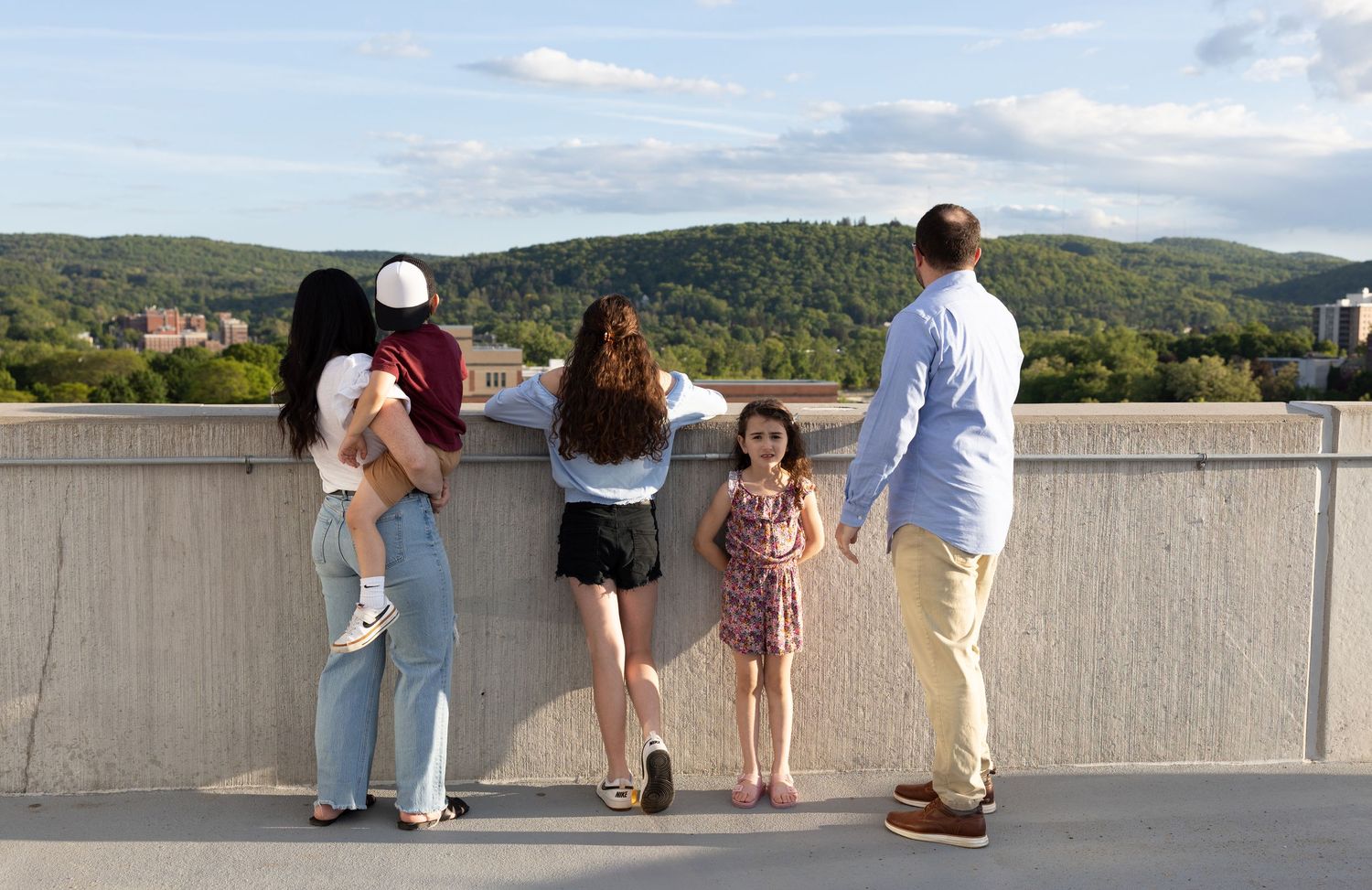 A group of people stand at a scenic overlook with rolling green hills and blue sky in the background.
