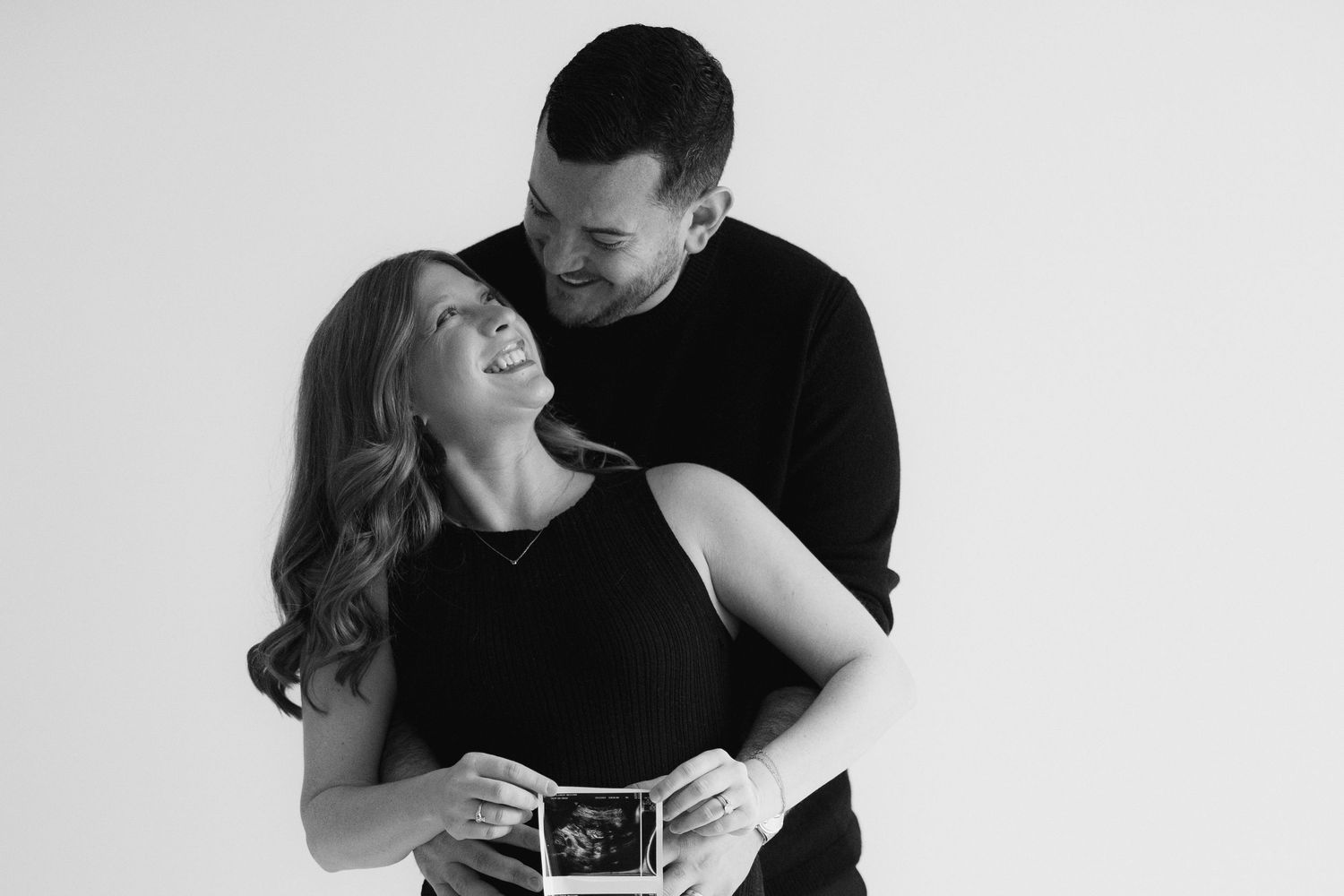 A couple holds an ultrasound photo while embracing in a black and white pregnancy announcement photo series.