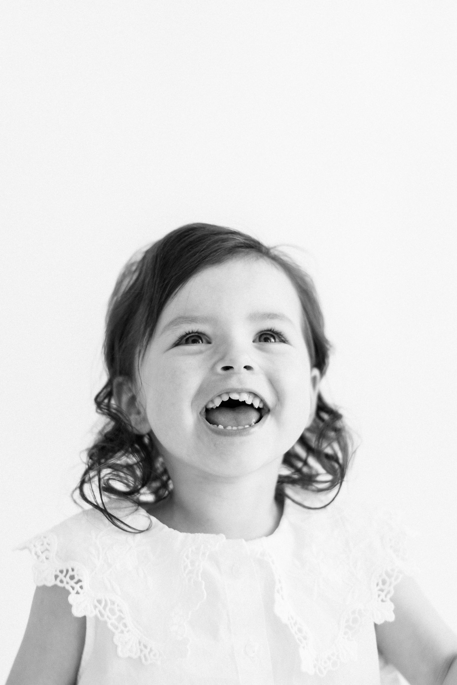 Black and white portrait of a joyful child with curly hair wearing a white lace dress and laughing with pure delight.
