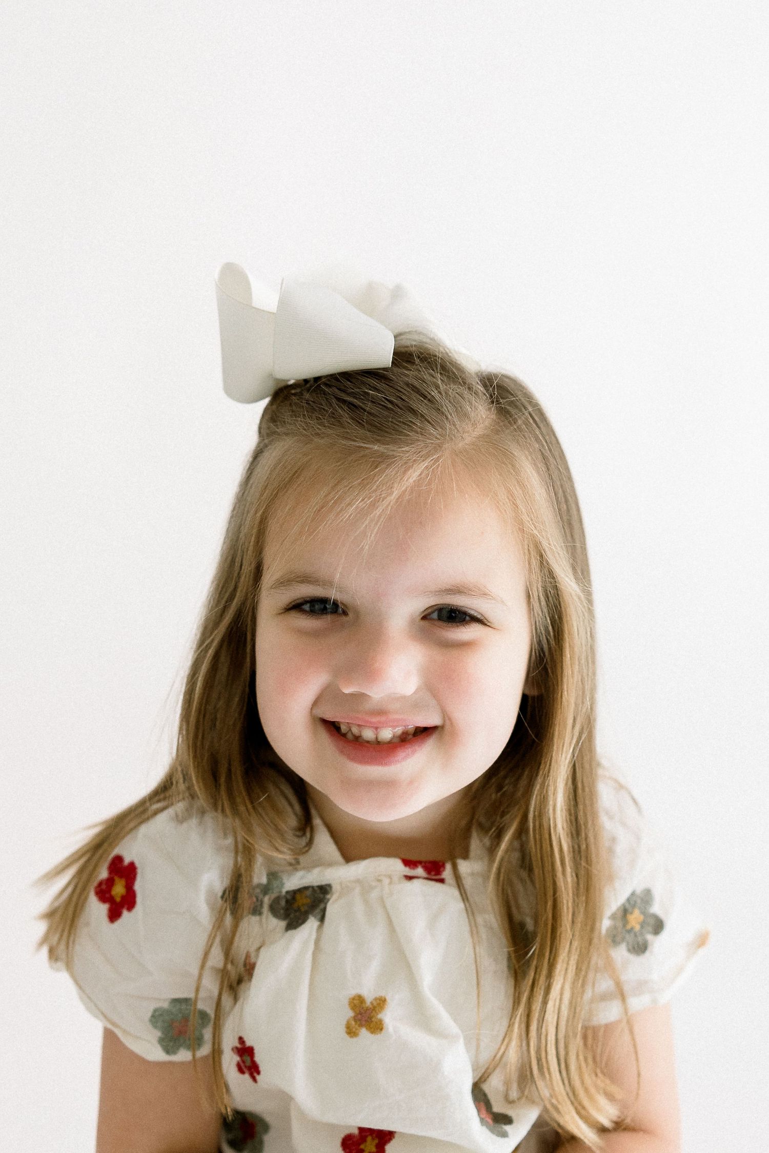 Child wearing a white floral dress and white bow smiles against a light background.