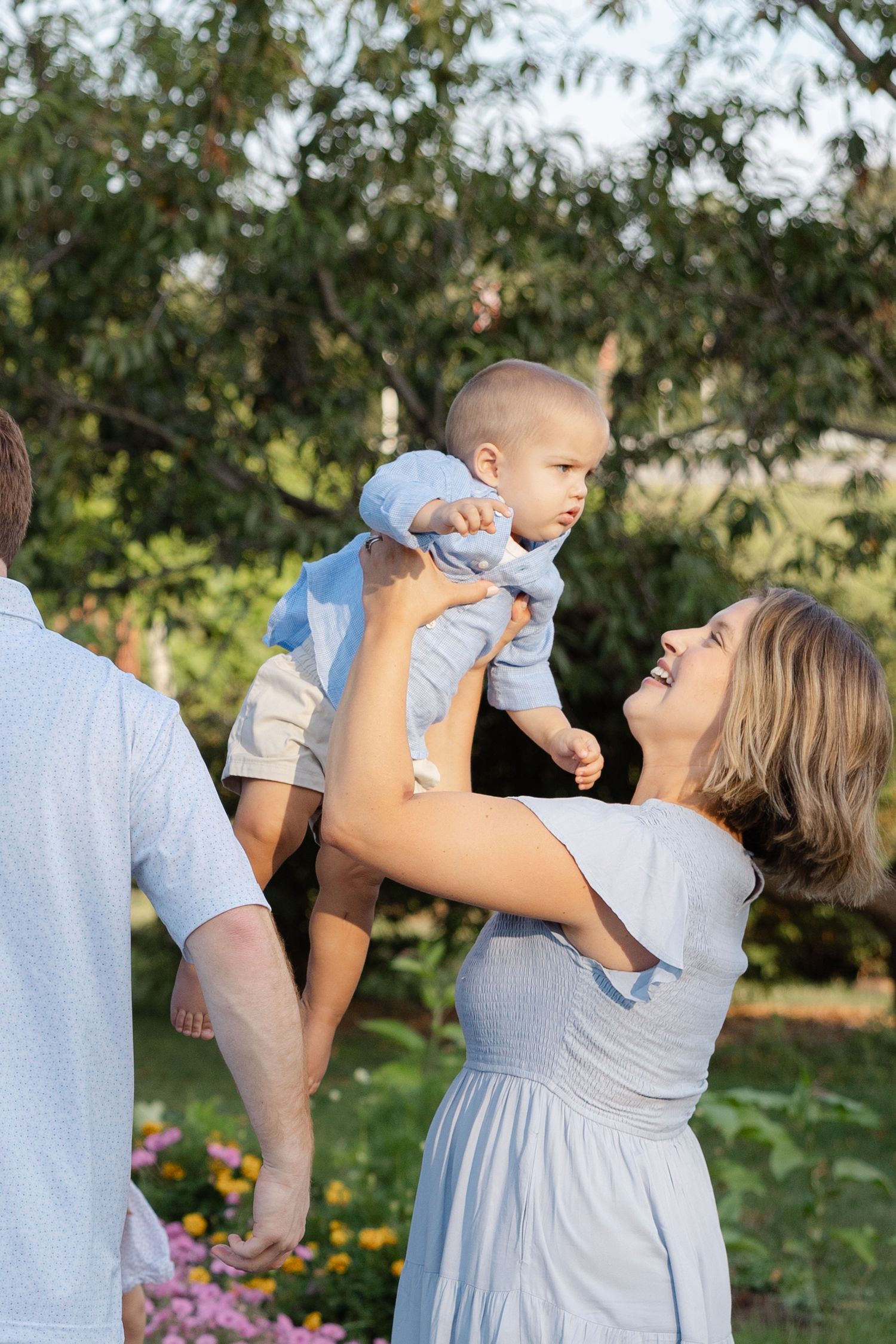 Joyful moment in a garden as a parent lifts a baby wearing light blue clothing against a natural backdrop of trees and flowers.