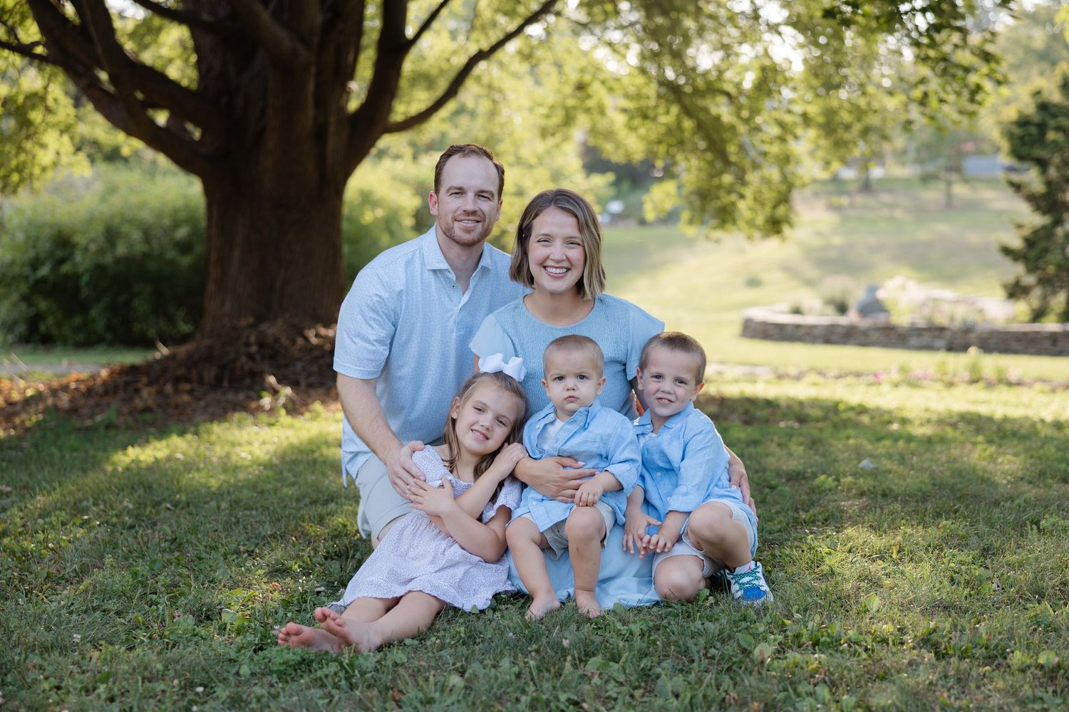 A family in coordinating light blue outfits sits together on grass under a shady tree at cutler botanic gardens in Binghamton ny
