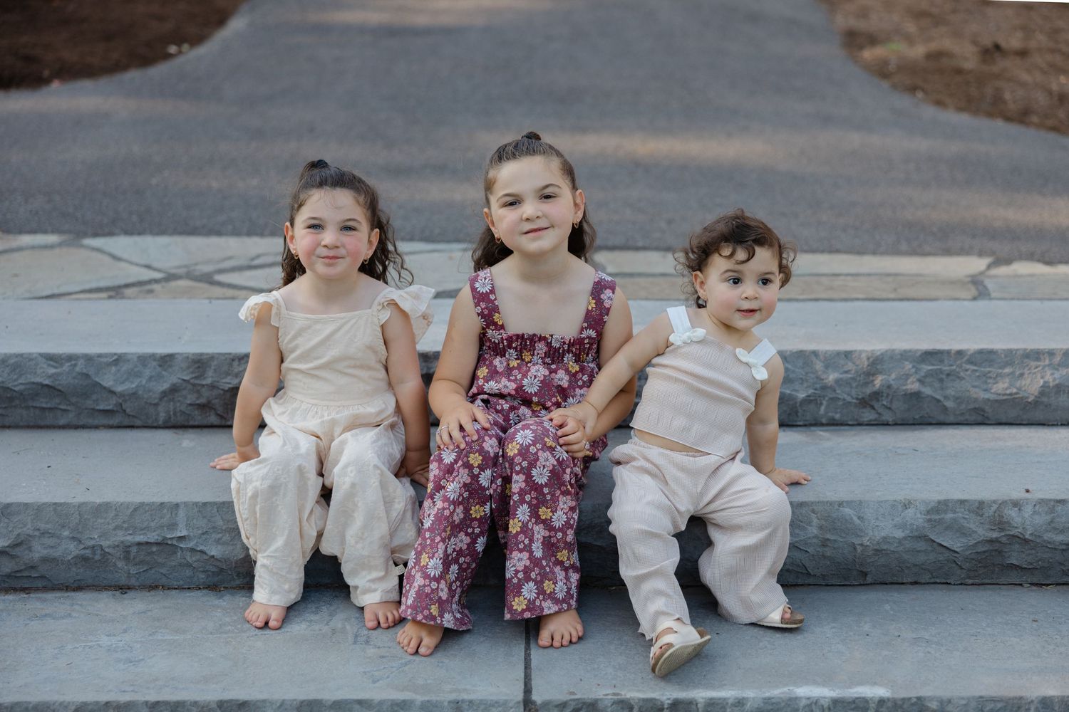 Young children sit together on outdoor concrete steps wearing casual summer outfits in matching shades of white and pink.