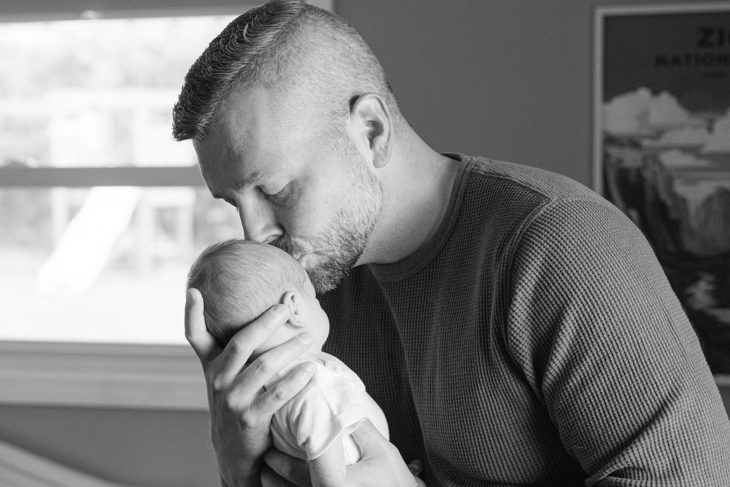 A tender black and white portrait of a parent gently kissing their newborn baby in a sunlit room.