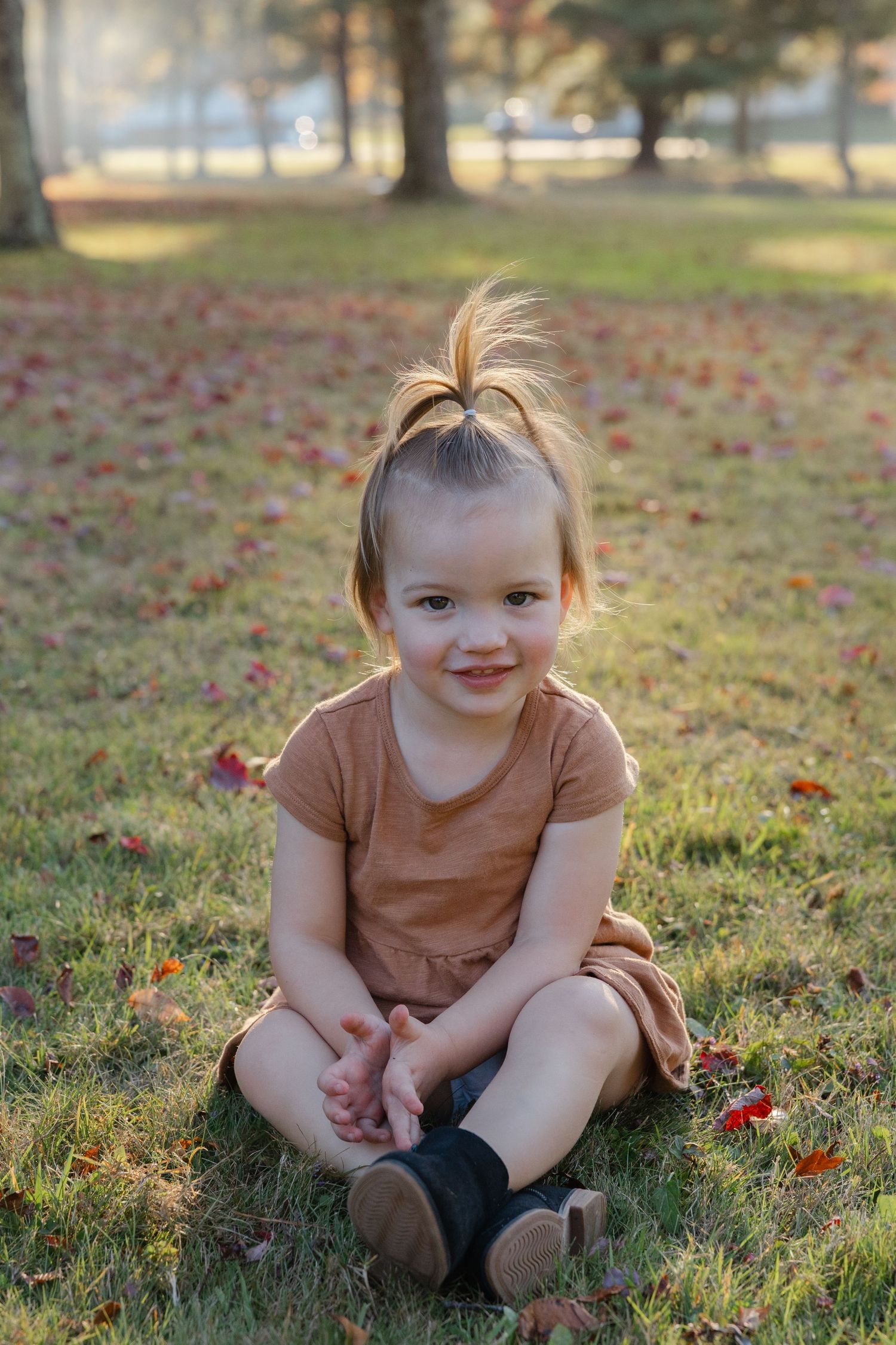 Small child in brown dress sits on grass with autumn leaves scattered around during golden hour.