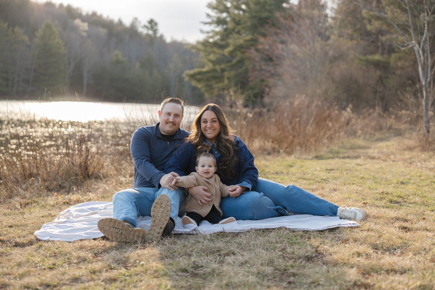 Family of three sitting on a blanket outdoors near a lake, smiling together on a sunny autumn day.