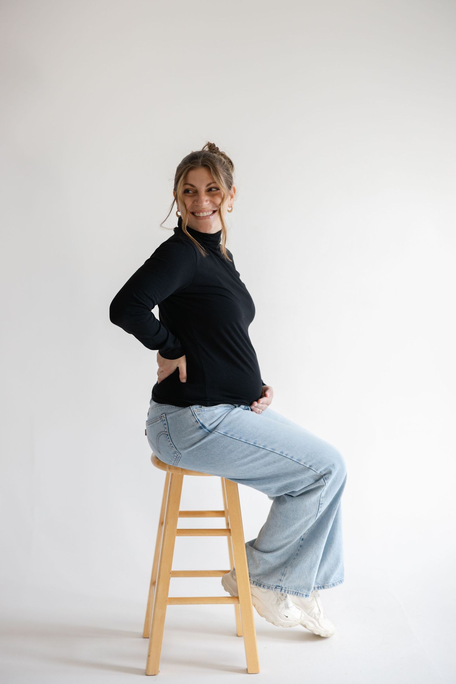 Someone in a black turtleneck sweater and light blue jeans sits casually on a wooden stool against a white backdrop.