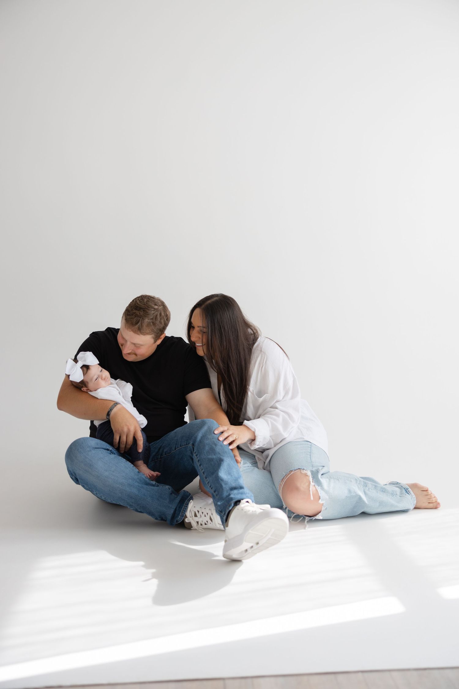 Couple in casual denim sits together on white floor while cuddling their small dog.