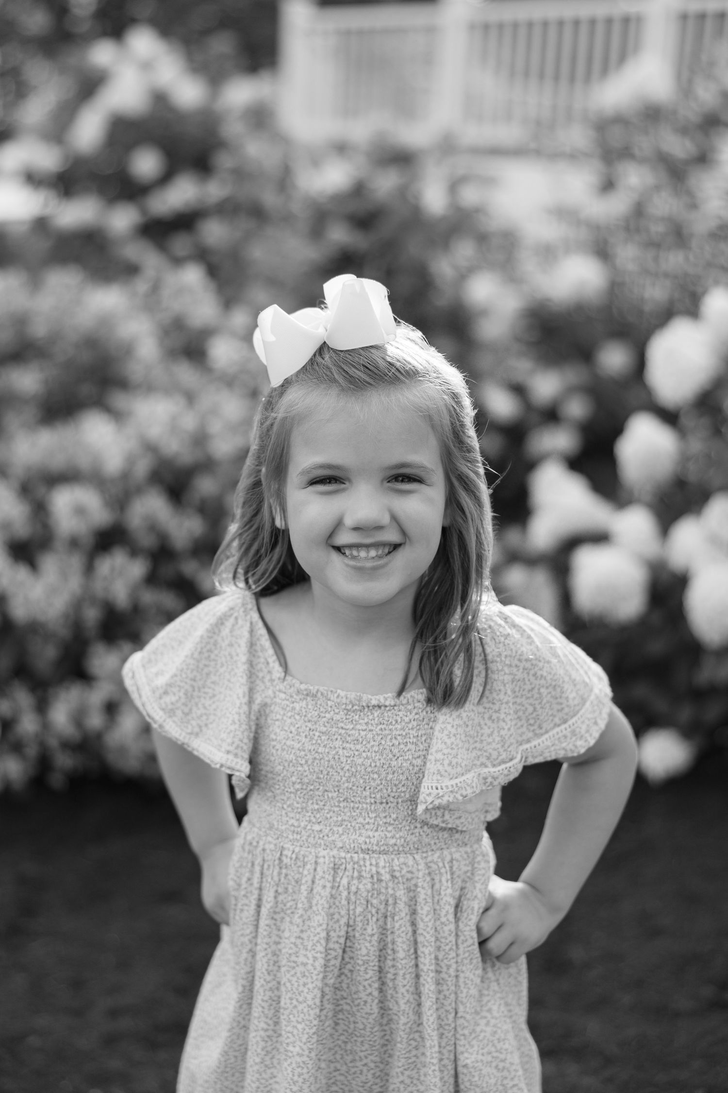 Black and white portrait of a young child wearing a white dress with a bow in their hair, smiling outdoors.