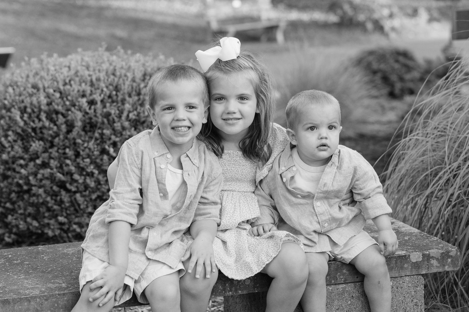 Black and white portrait of three young children sitting together on a bench in garden setting.
