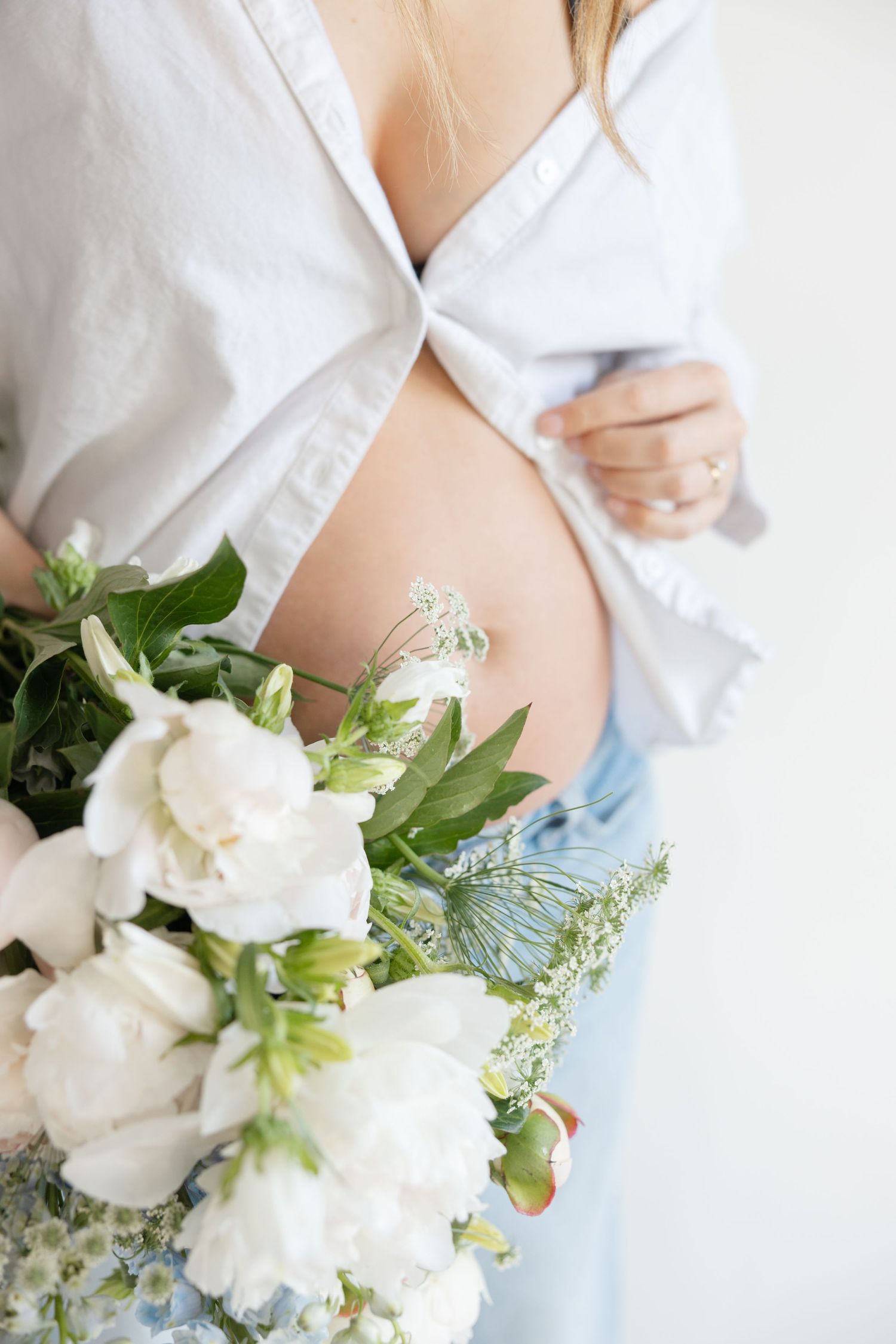 A delicate white floral bouquet rests against a pregnant belly draped in soft white fabric and blue denim.