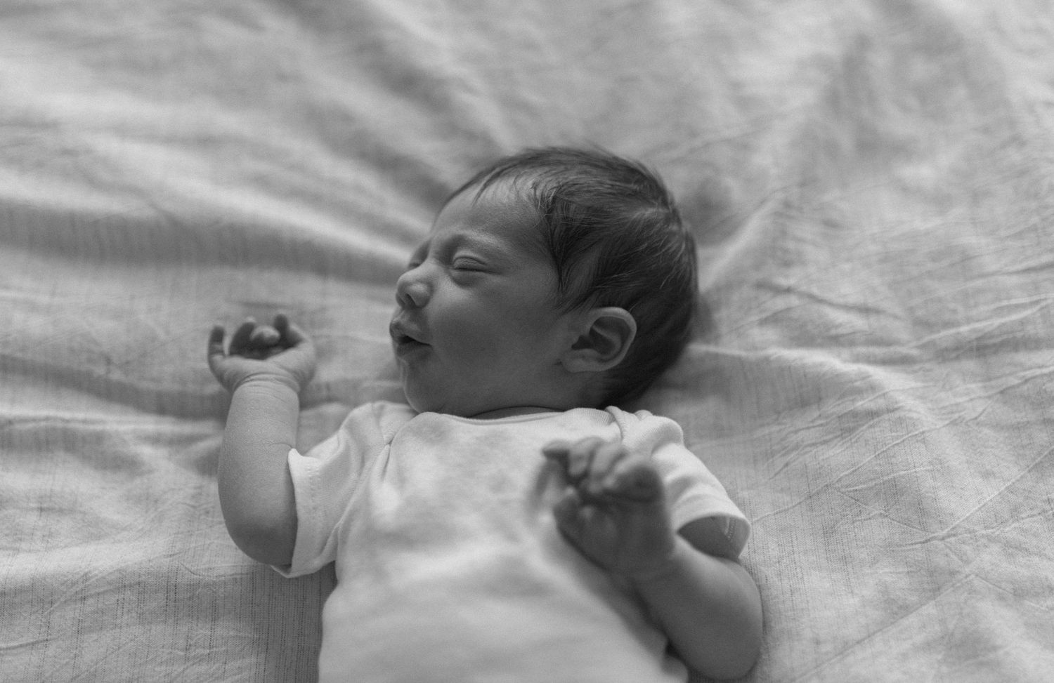 Newborn baby sleeping peacefully on soft white textured fabric in black and white photograph.