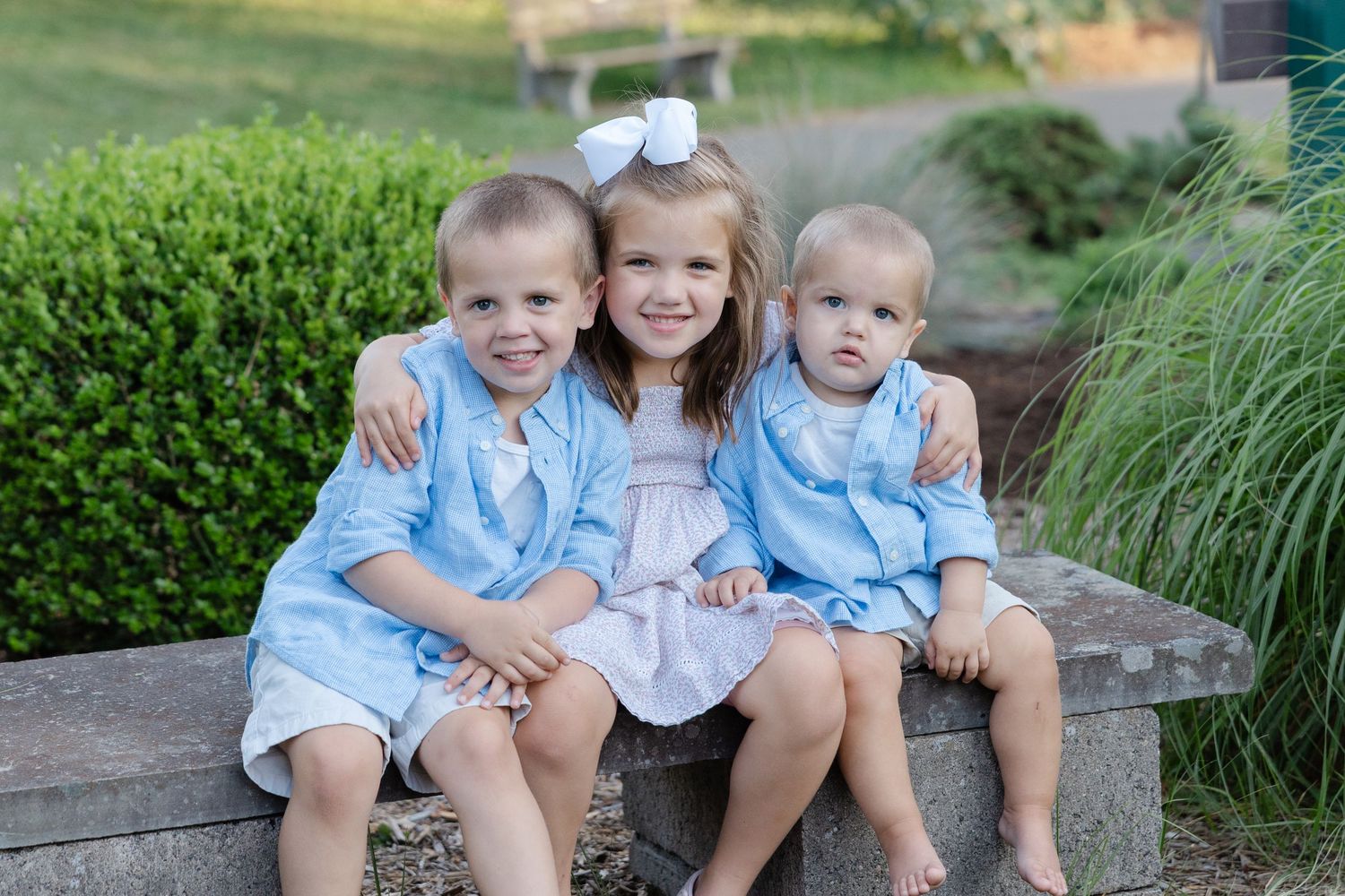 Three children in coordinating light blue and white outfits sit together on a wooden bench in a garden setting.