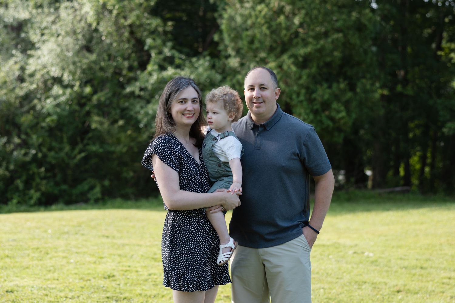 A family stands together on a grassy lawn surrounded by trees on a sunny day.