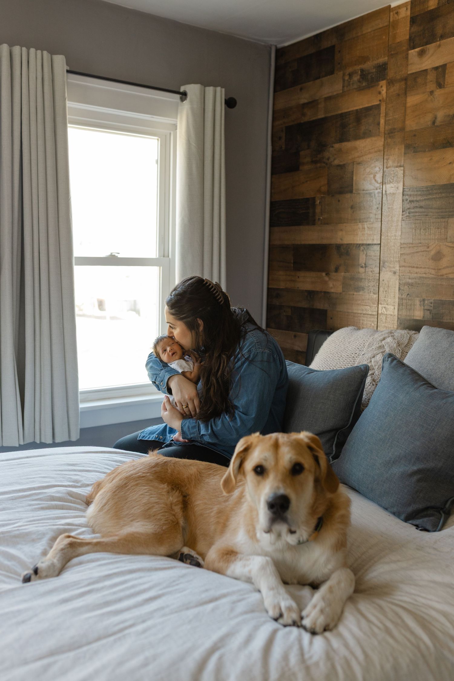 Baby rests against parent on couch while golden retriever relaxes on white bedding in cozy room with wooden accent wall.
