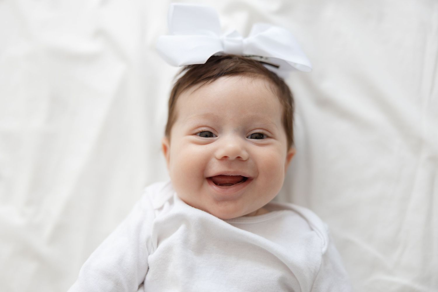 A young baby wearing a white onesie laying on a white blanket with a joyful smile.