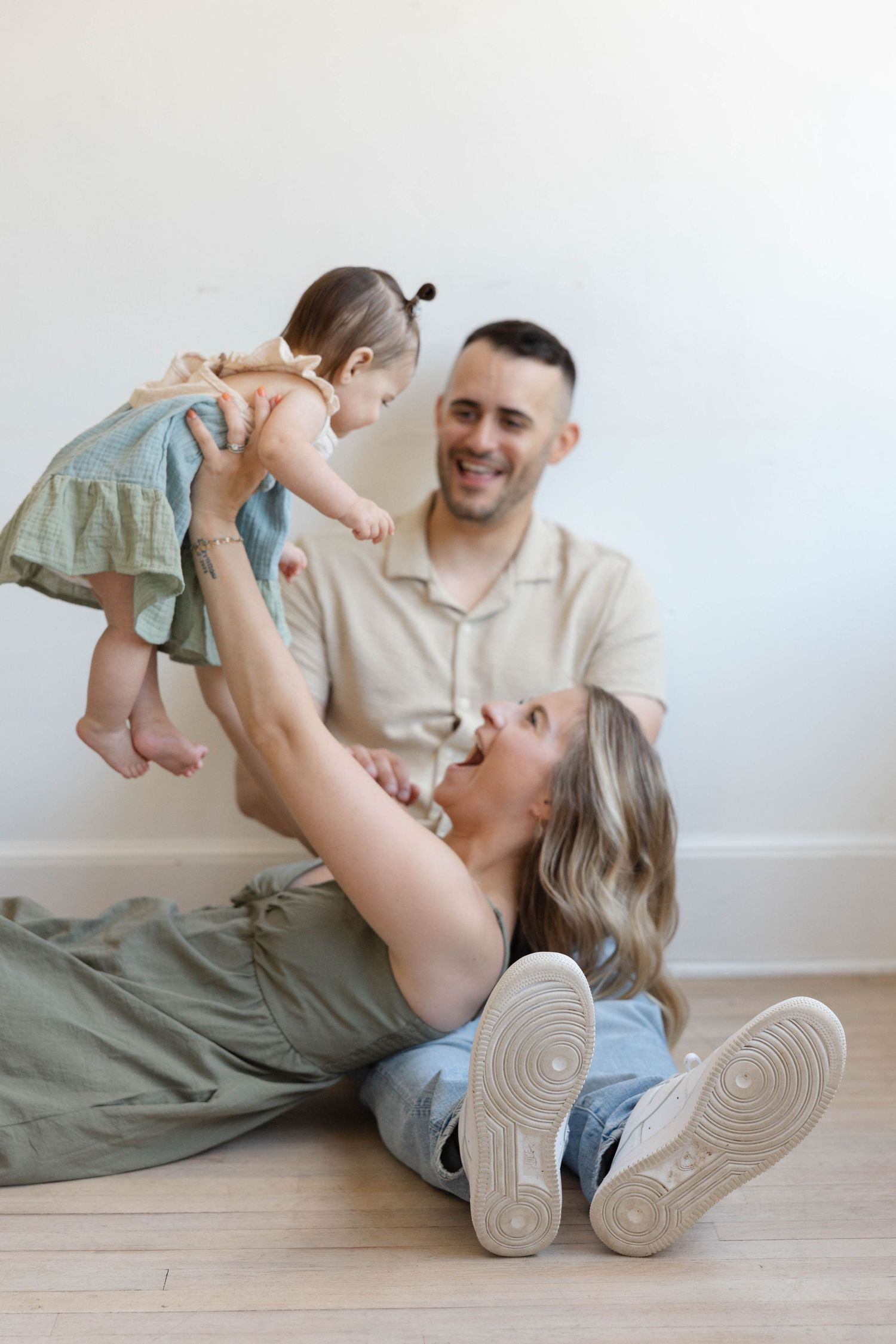 Happy family with mom lifting baby girl while dad smiles, sitting on floor against white wall.