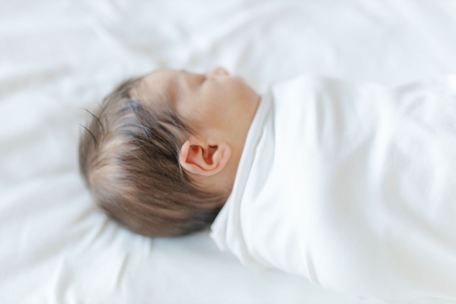 Sweet sleeping baby wearing white onesie rests peacefully on white bedding with soft natural lighting.
