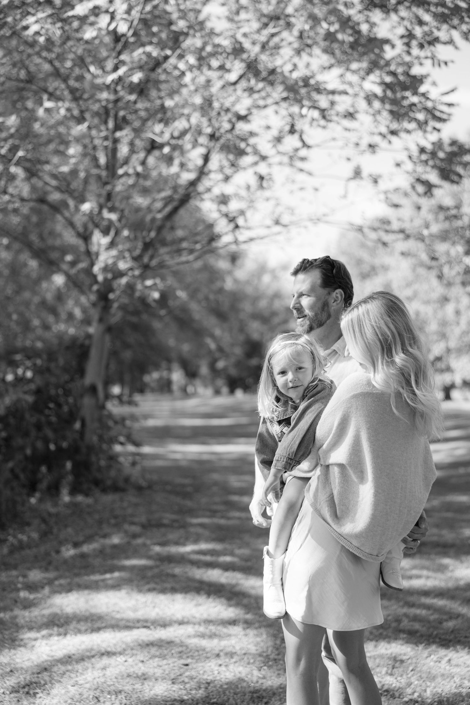 Black and white photo of a family with a young child enjoying a peaceful walk through a tree-lined park path.