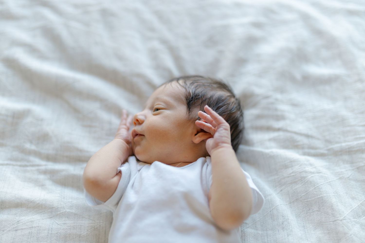 Peaceful newborn baby in white onesie sleeping on soft white bedding with hands raised near face.