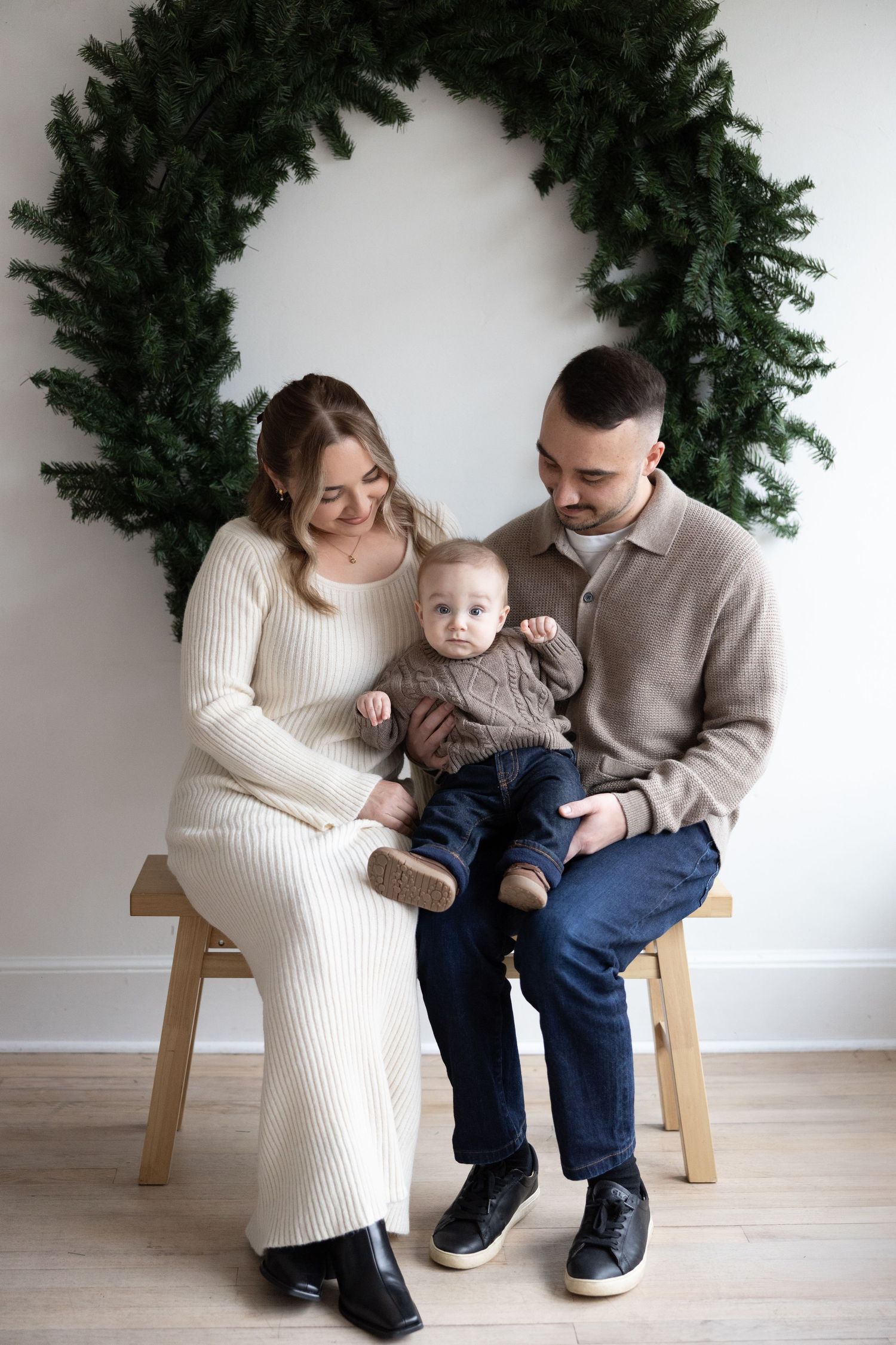 A family in cream and beige sweaters sits together on a wooden bench against a white wall with greenery.