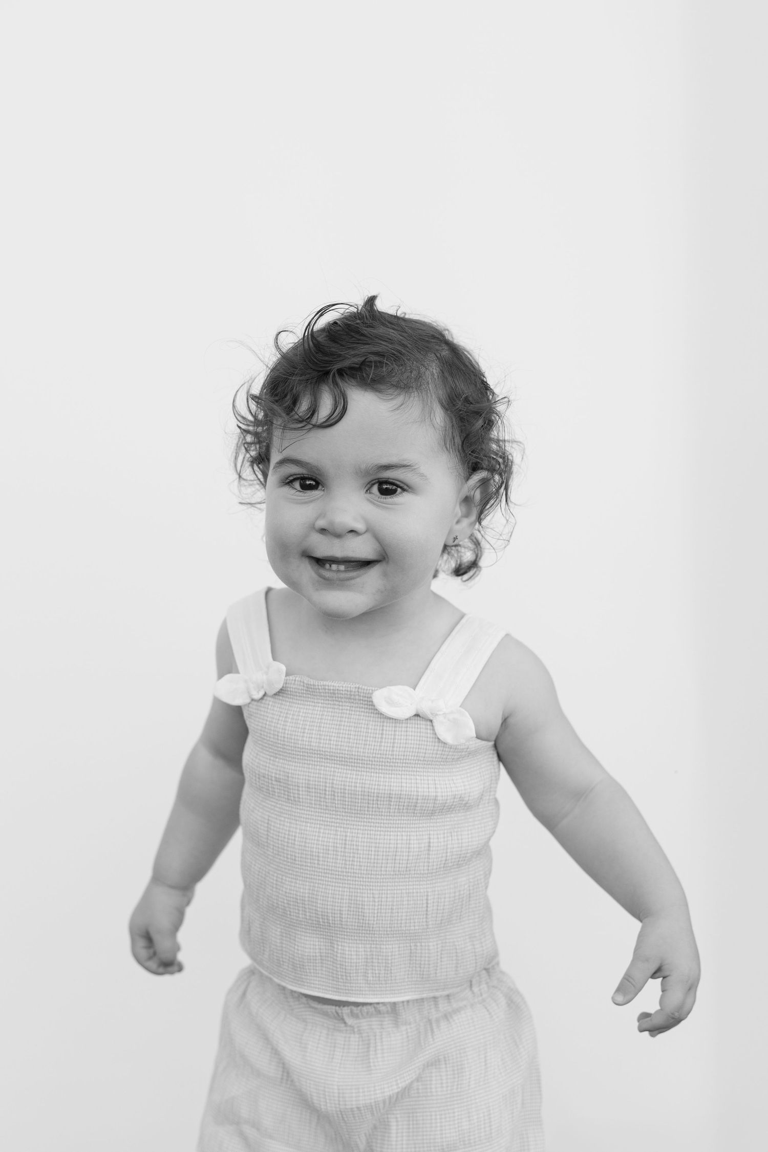 A happy toddler wearing a white ribbed tank top with bow details smiles while playing against a light background.