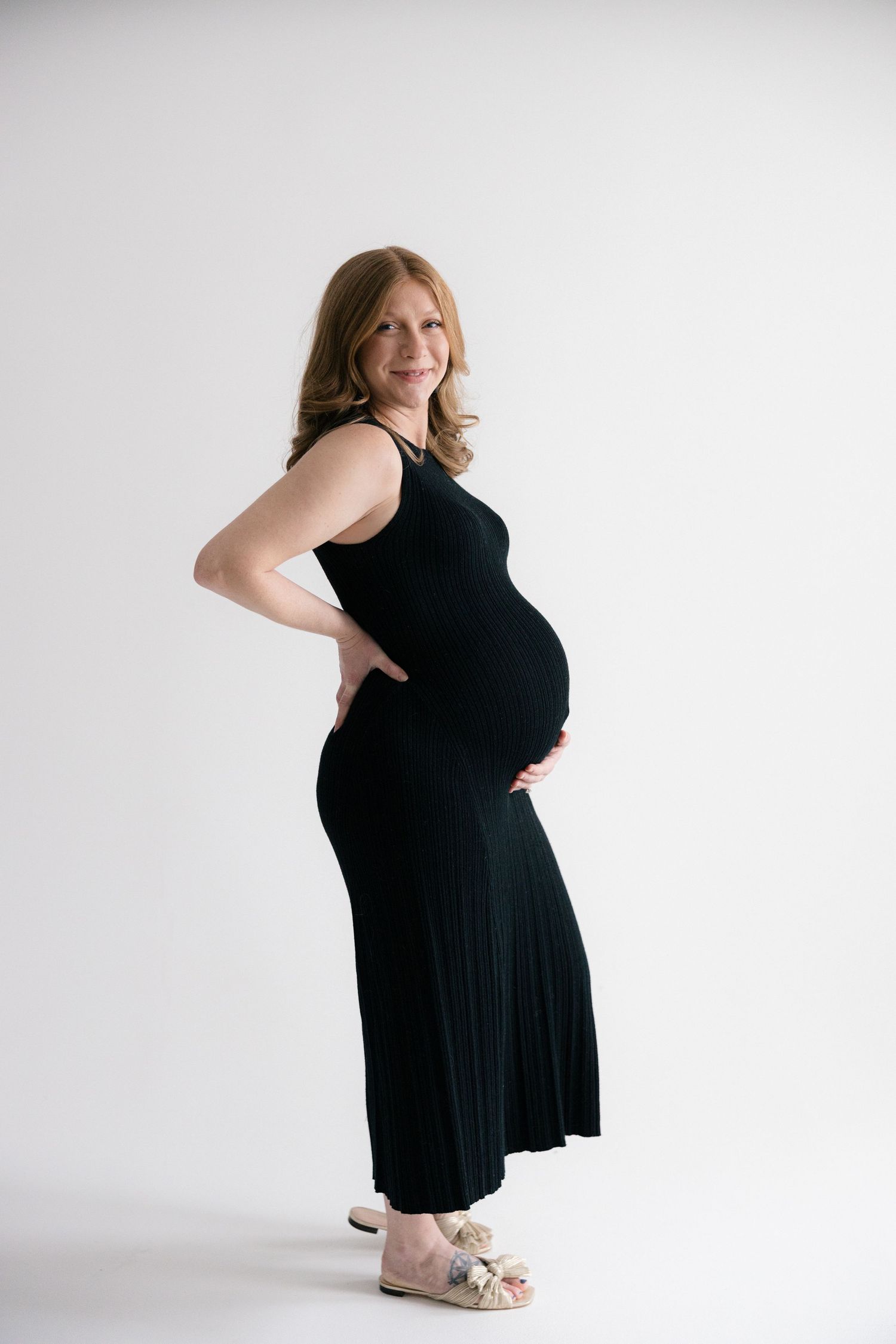 A pregnant woman in a black maxi dress poses sideways against a white backdrop, showing her baby bump.