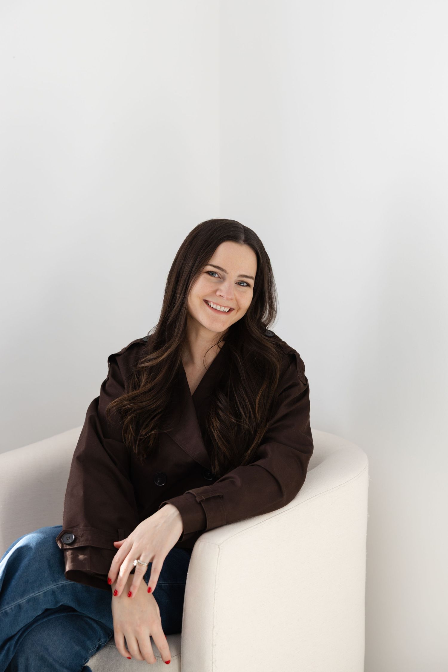 Smiling brunette woman in brown jacket and jeans sitting relaxed in white chair against light background.