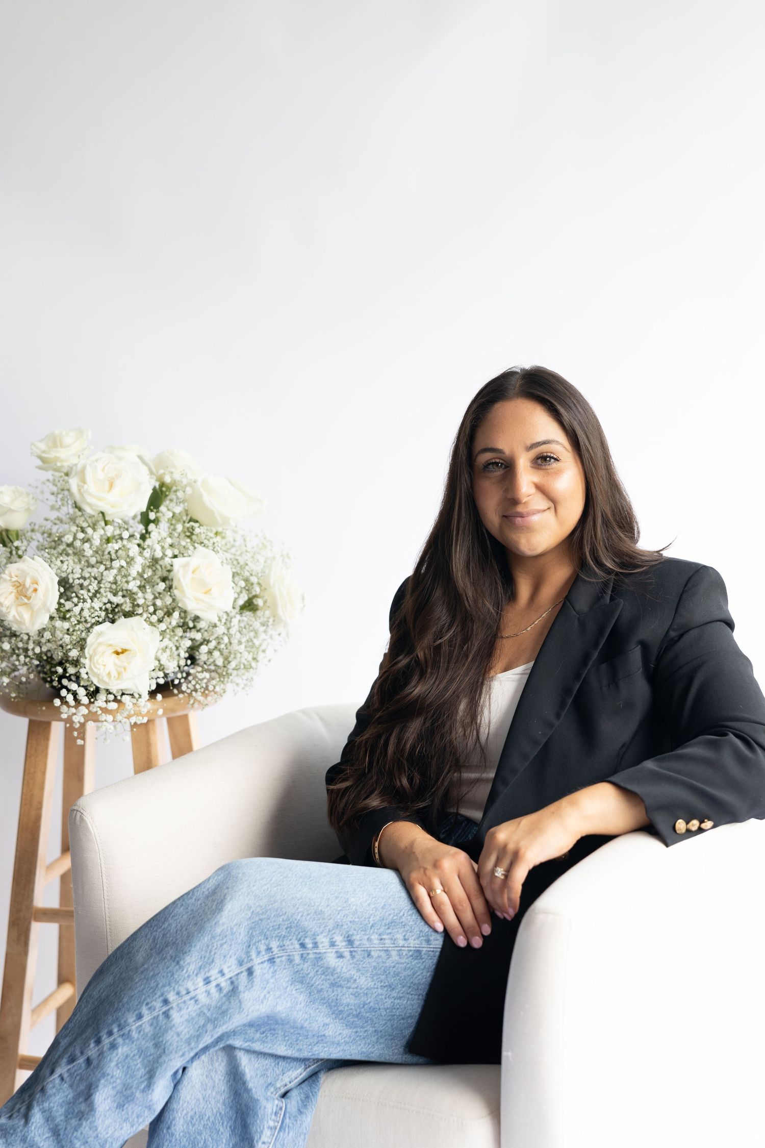 Professional portrait of someone relaxing on a white couch wearing a black blazer near white floral arrangement.
