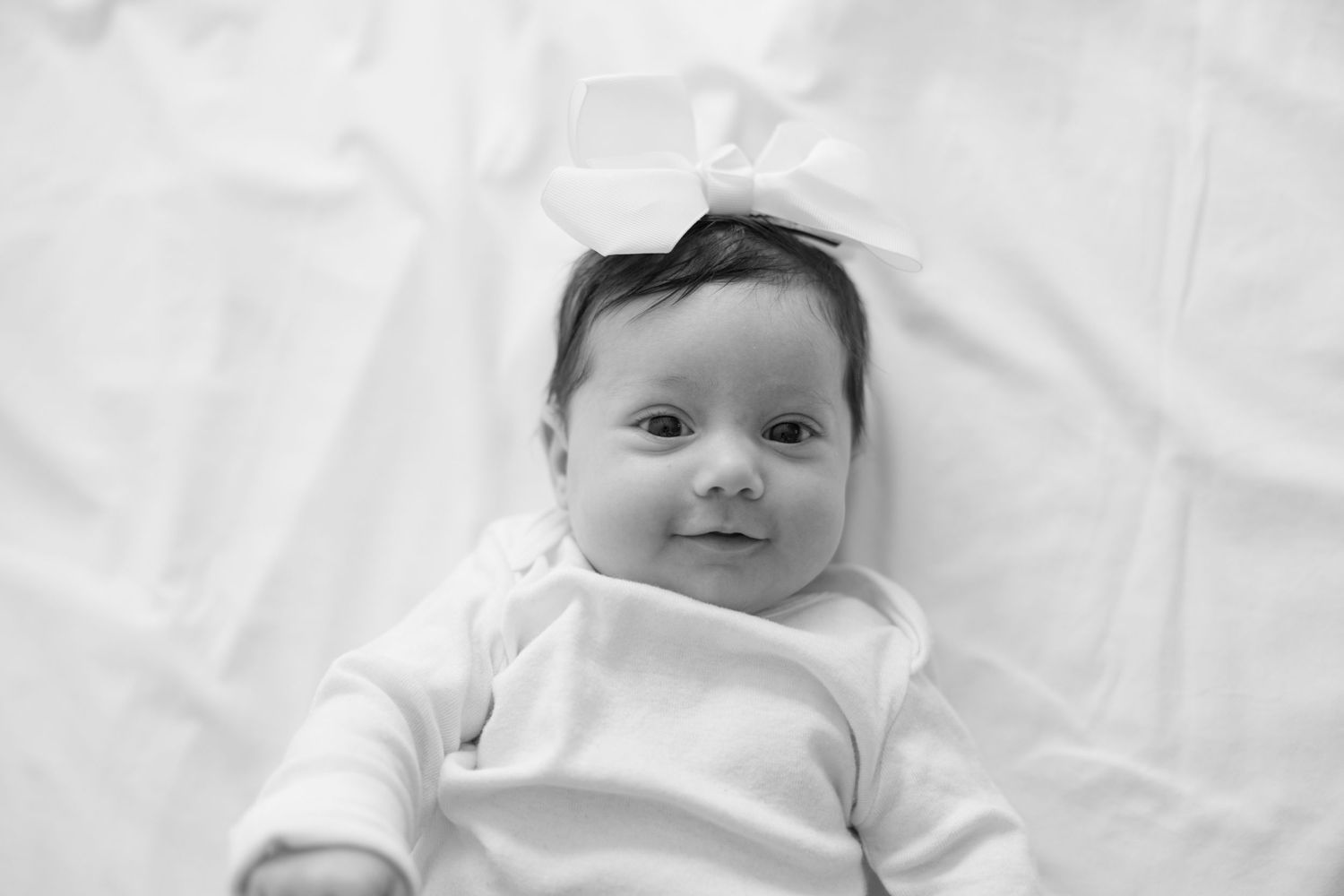 A smiling baby with dark hair lies on white bedding wearing a light-colored outfit.