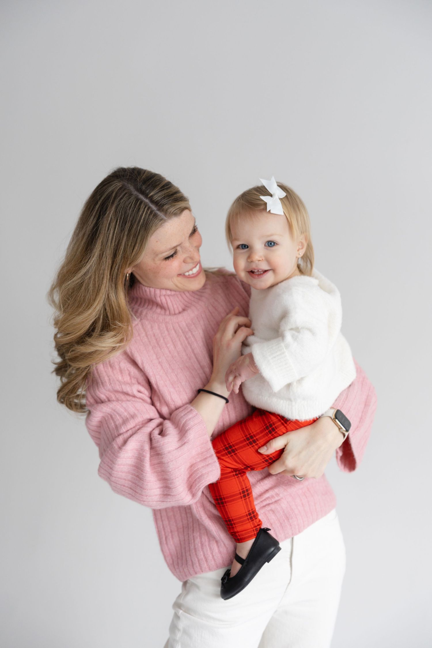 A tender moment captured against a gray backdrop showing warm winter fashion with pink sweater and red plaid pants.