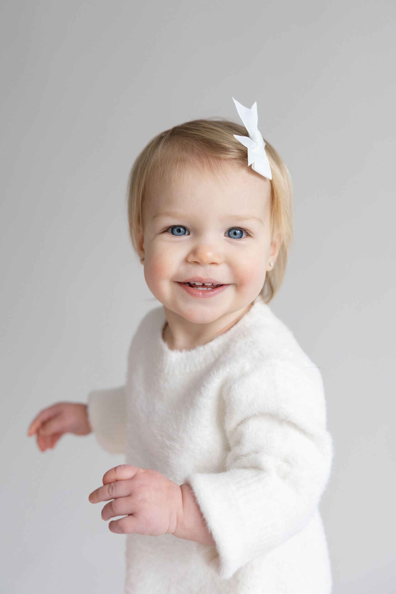 Toddler wearing white sweater and hair bow smiles against light gray background.