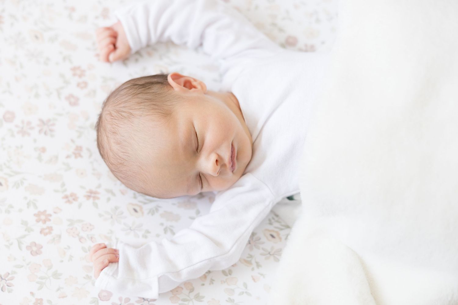 A newborn baby sleeps peacefully on white bedding surrounded by delicate floral patterns.