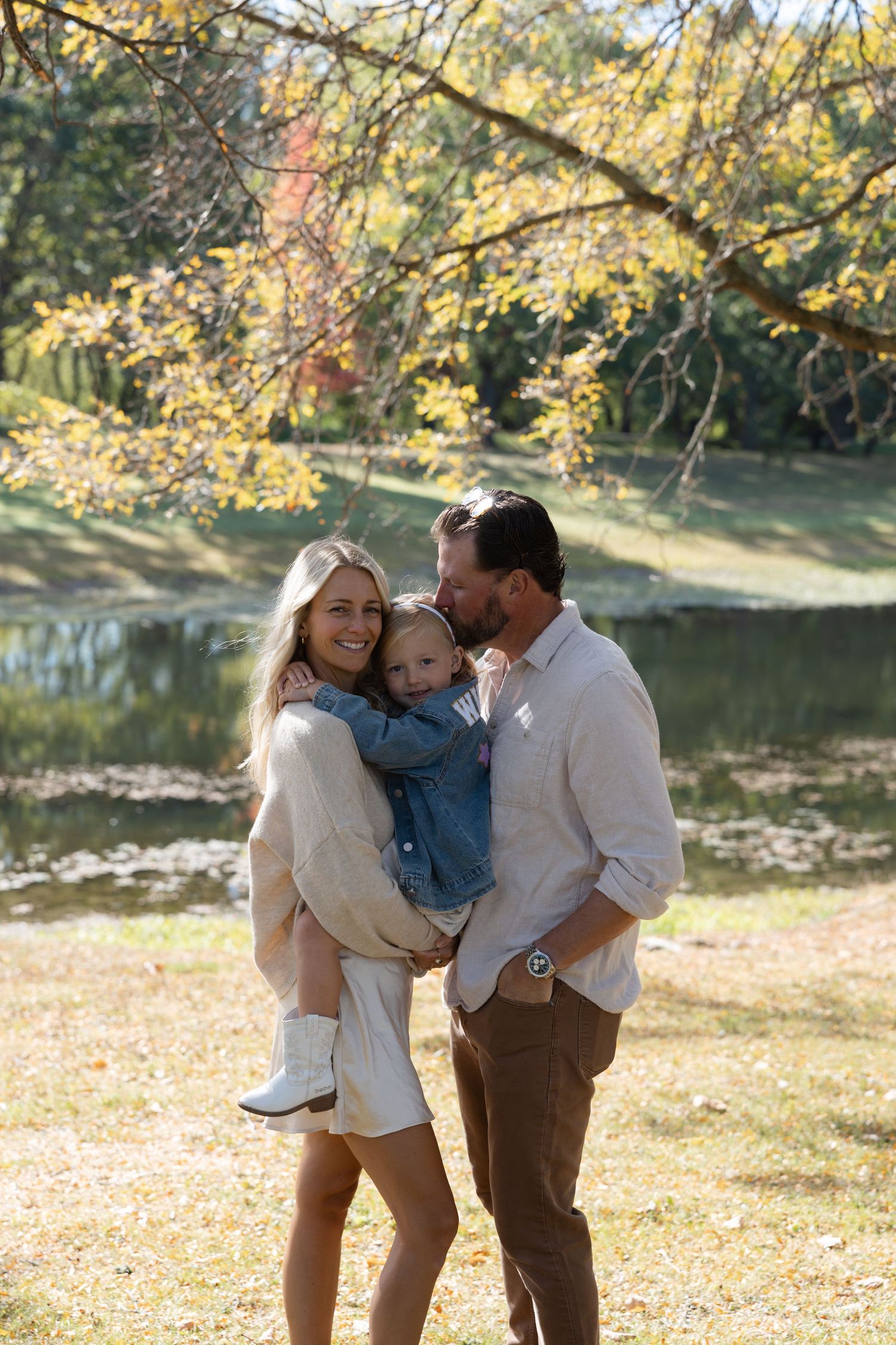A family shares tender moments together in a scenic autumn setting by a lake with golden foliage in the background.