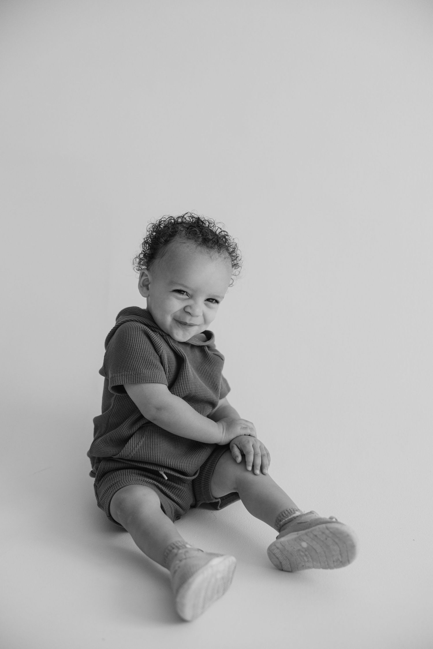 Black and white portrait series showing a baby in dark clothing sitting and curled up against a plain background.