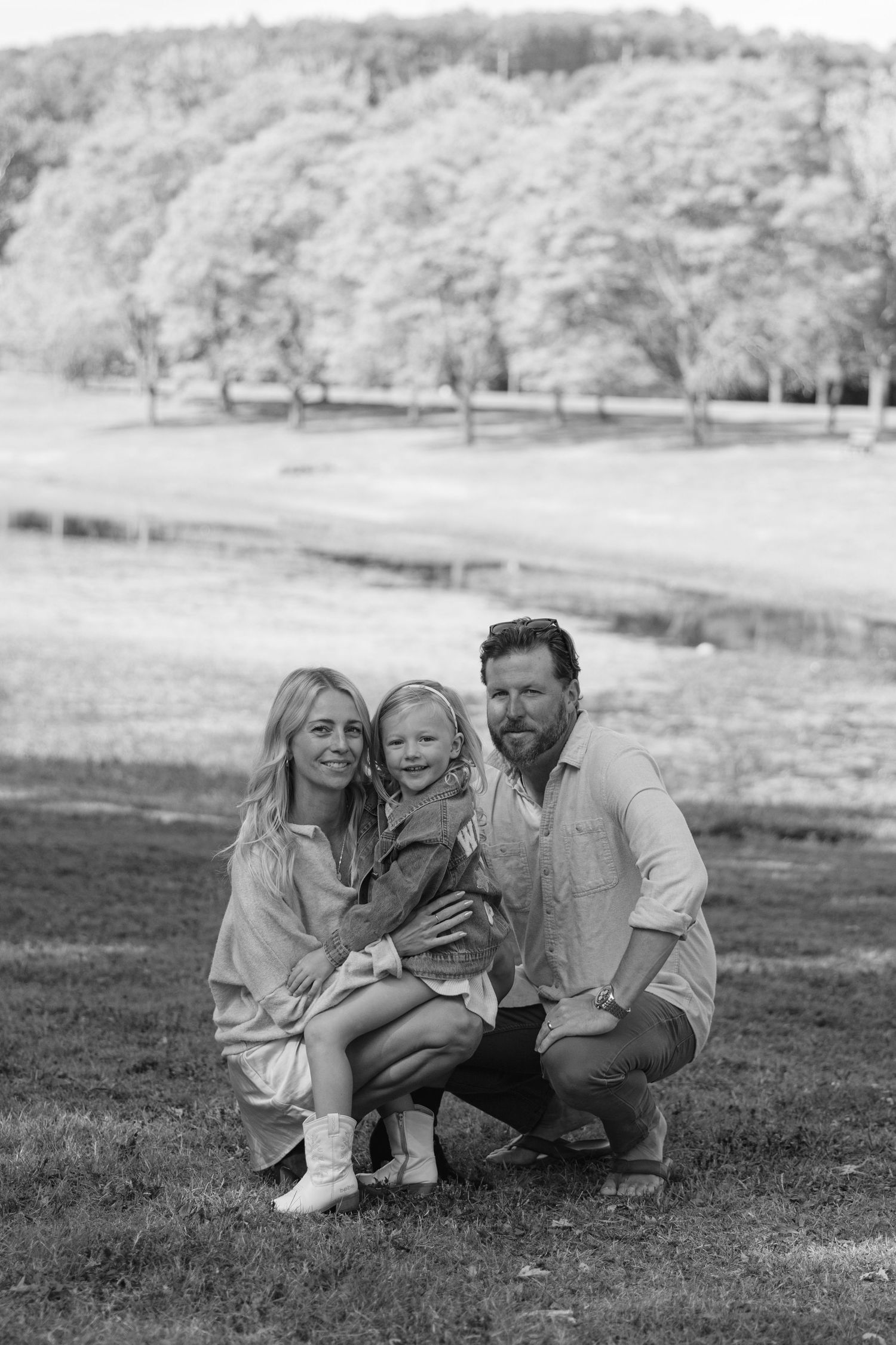 Black and white outdoor photo of family lying together on grassy hillside in natural setting.