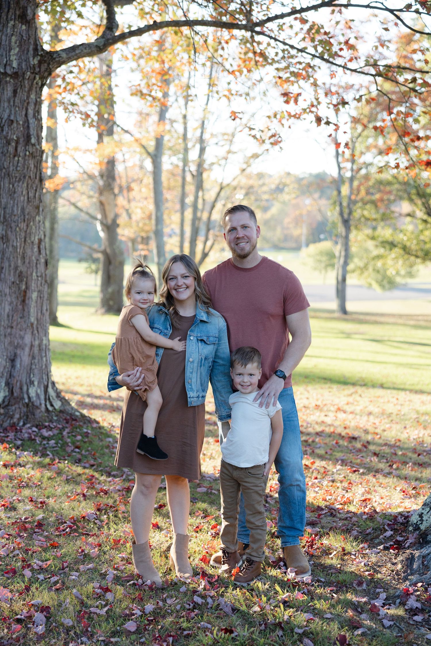 A family poses together in an autumn forest setting with colorful fallen leaves scattered on the ground.