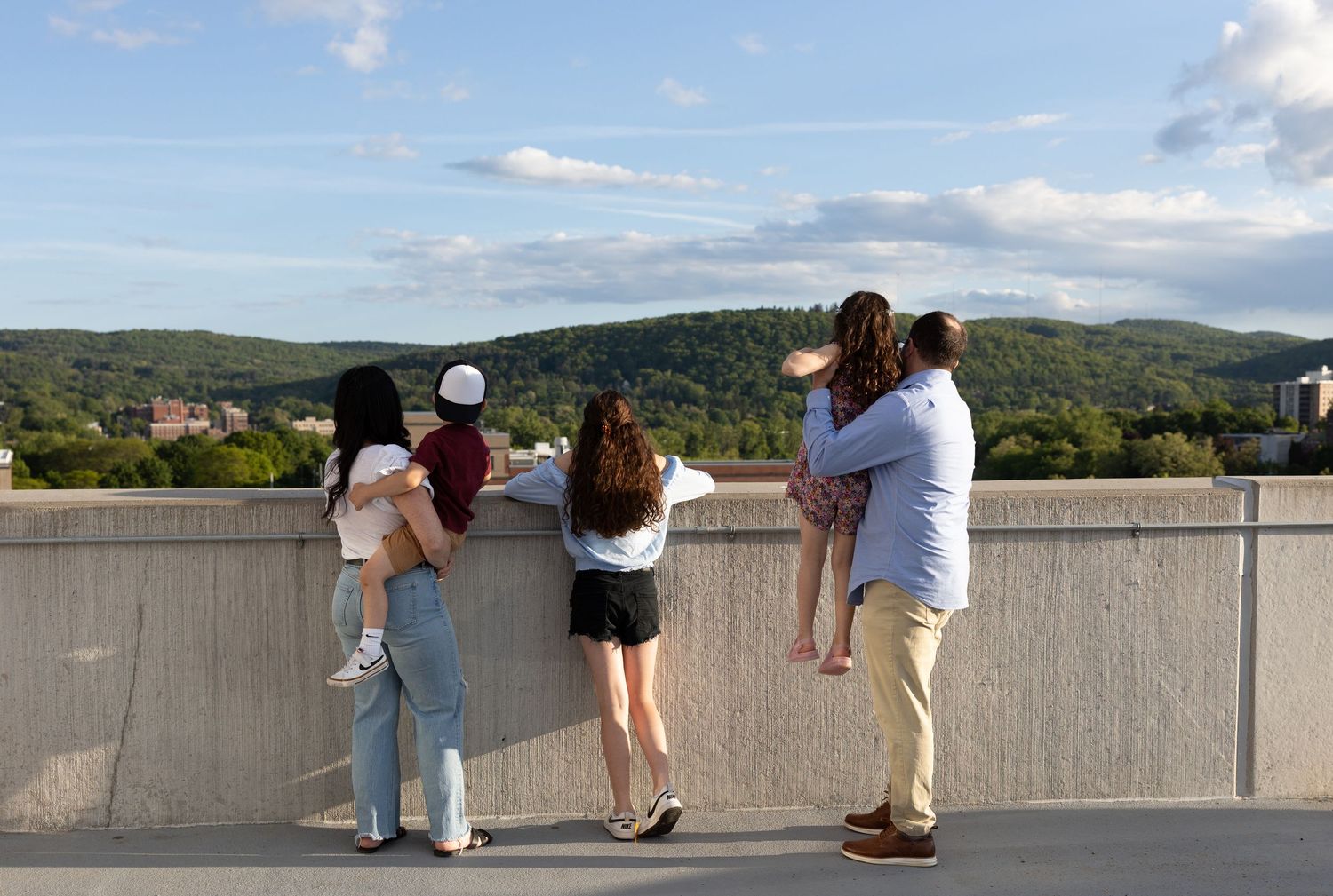 People look out from a concrete observation deck overlooking a scenic mountain landscape on a sunny day.