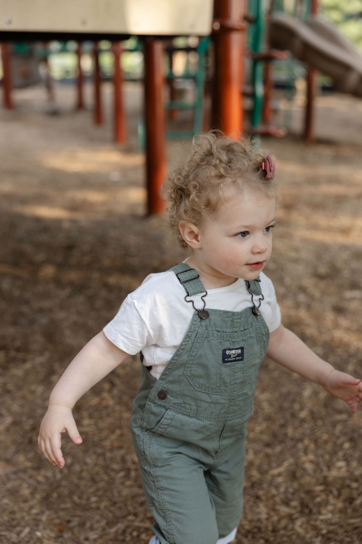 A toddler wearing olive green overalls and white shirt playing on wood chips under playground equipment at a park.