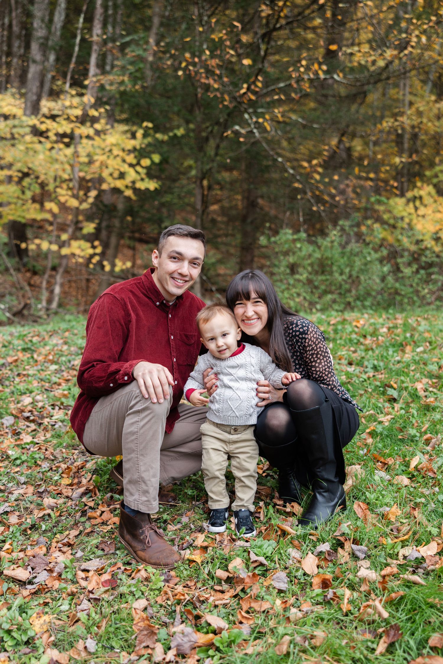 A family sits together on fallen autumn leaves in a wooded area wearing burgundy and dark colored clothing.