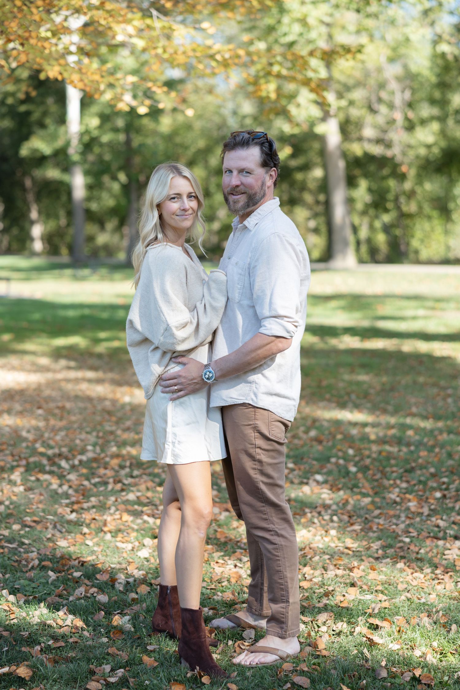 A couple embraces while standing in a sunlit autumn park with fallen leaves scattered on the ground.