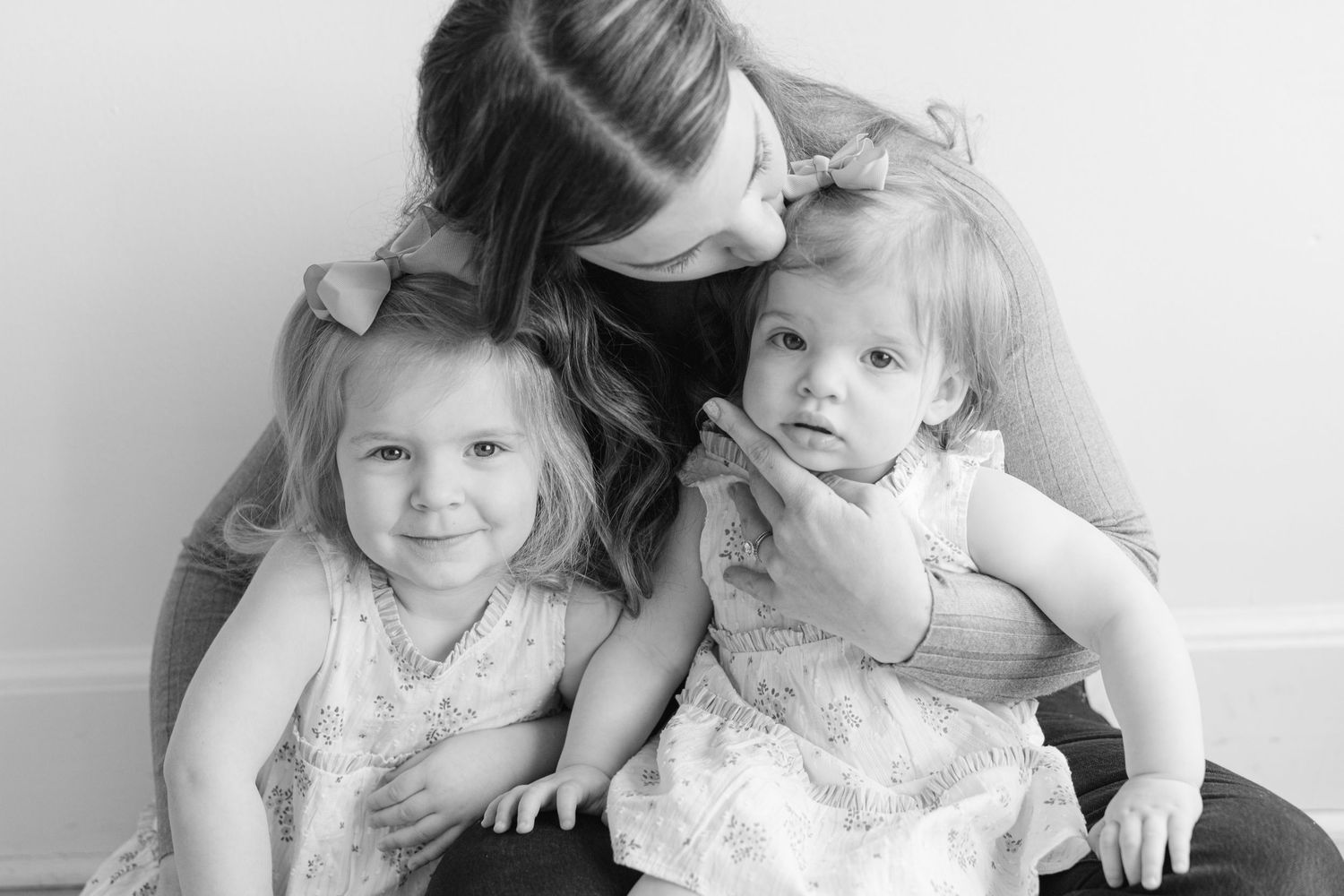 Black and white photo of a mother lovingly embracing her two young daughters in white dresses.