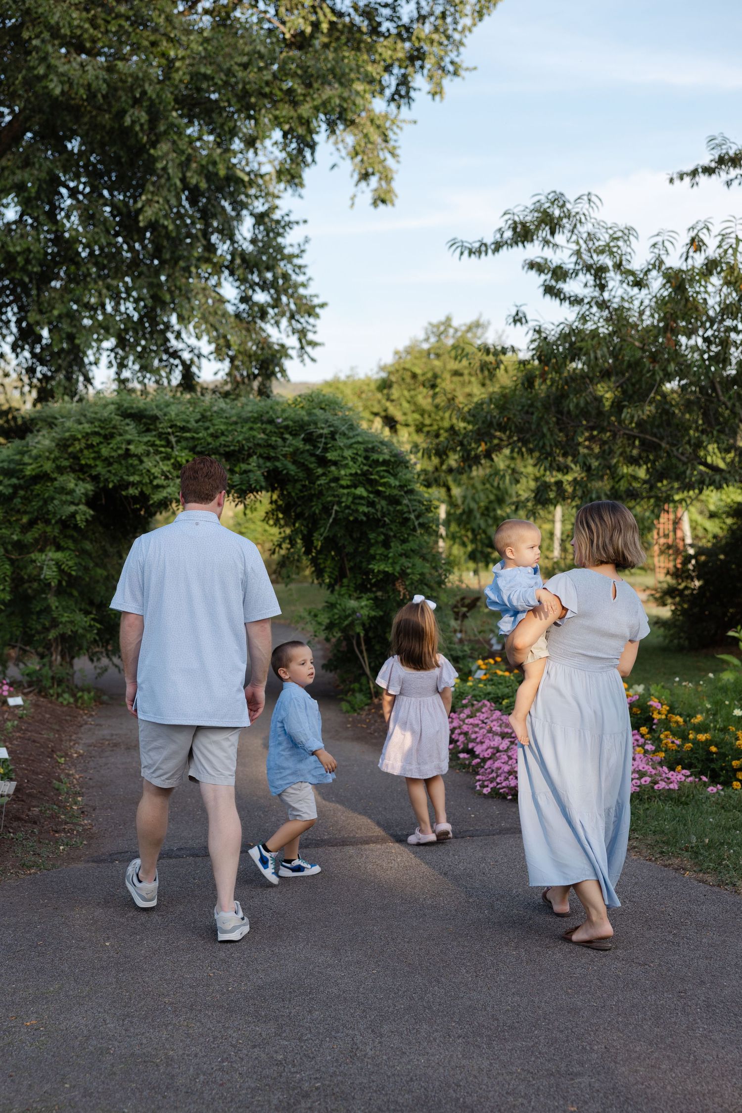 A group walks together on a garden path surrounded by flowers and trees on a sunny summer day.