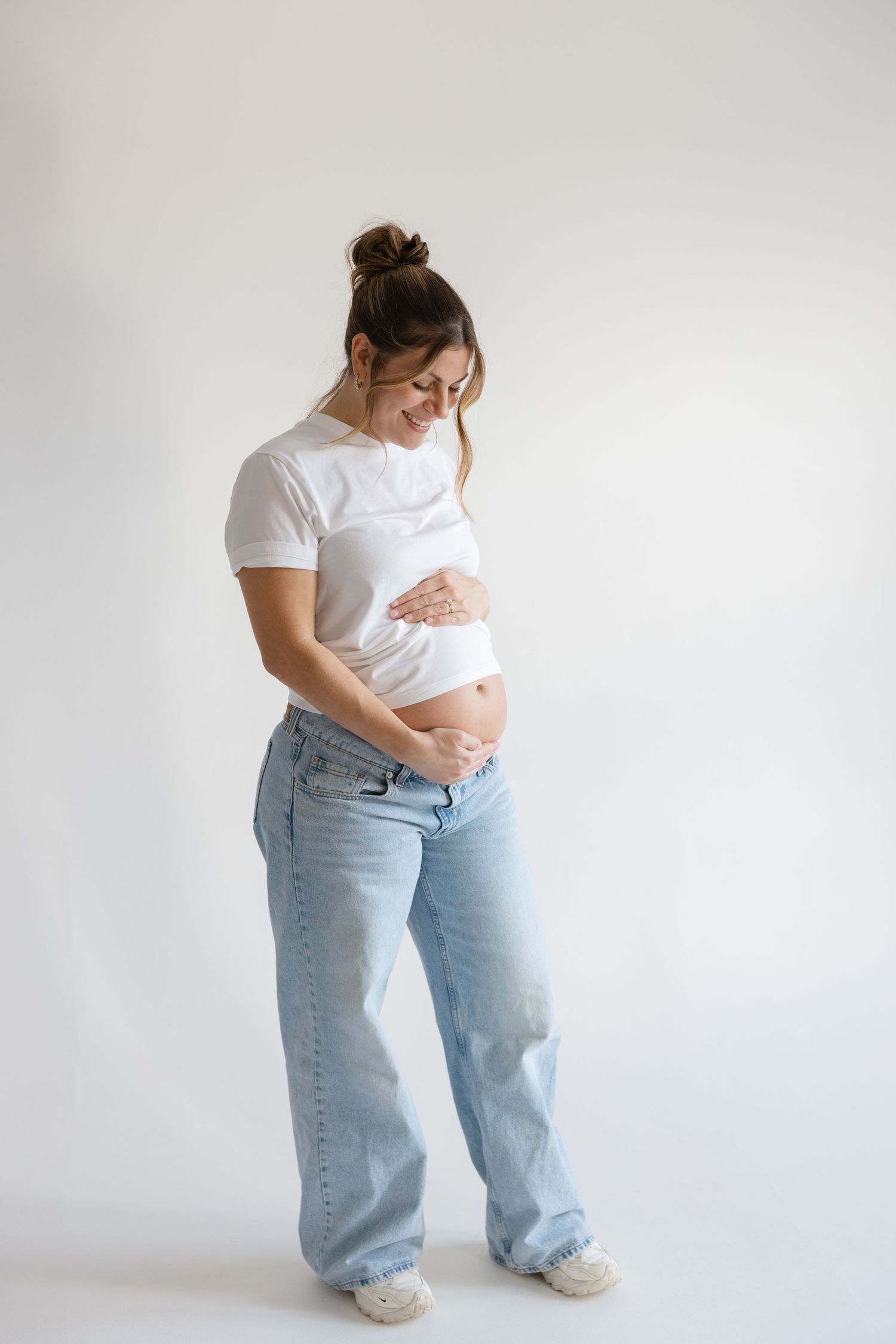 Pregnant woman in white t-shirt and baggy jeans poses against white background showing her growing belly.