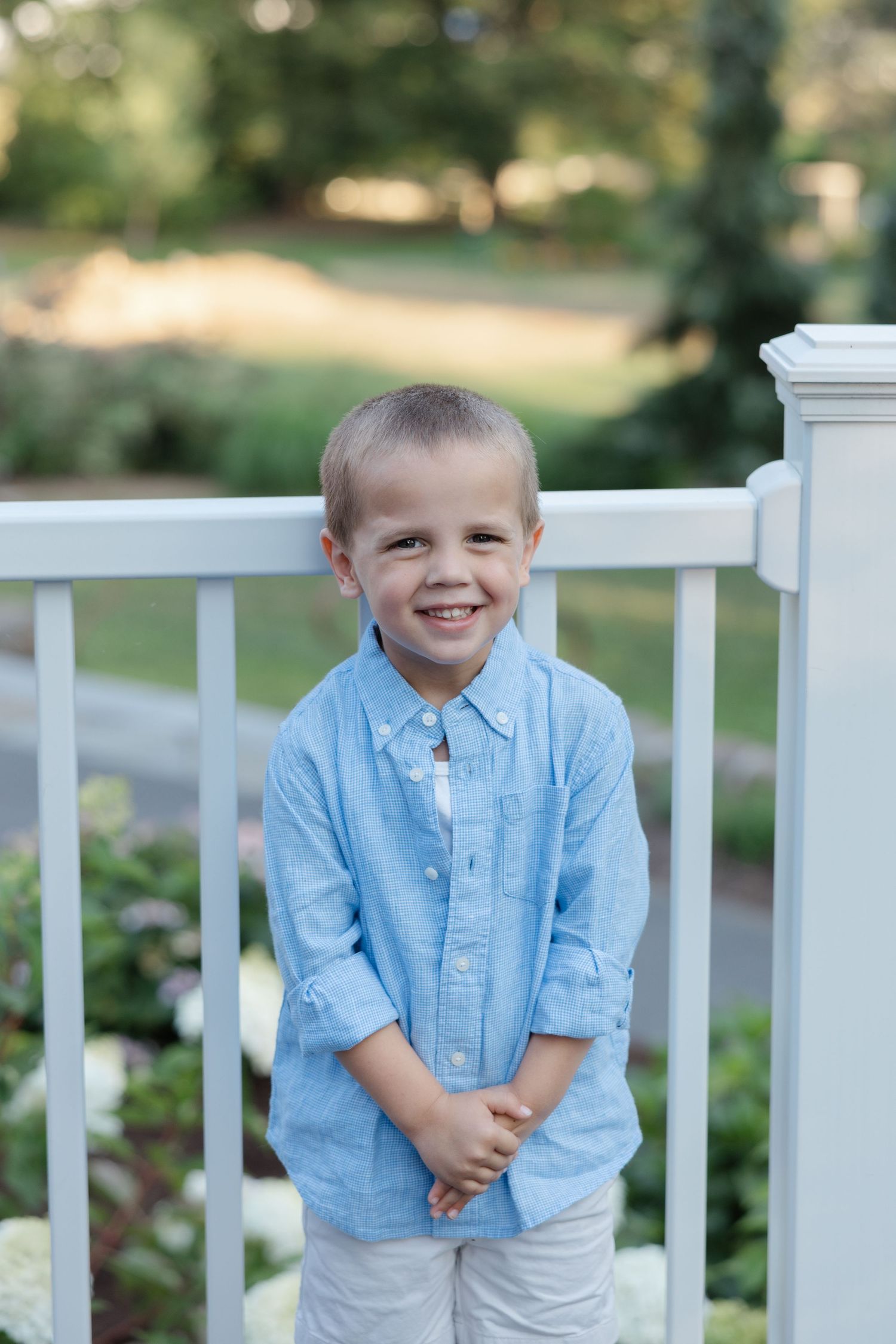 Child in light blue shirt standing by white porch railing outdoors with a bright smile.