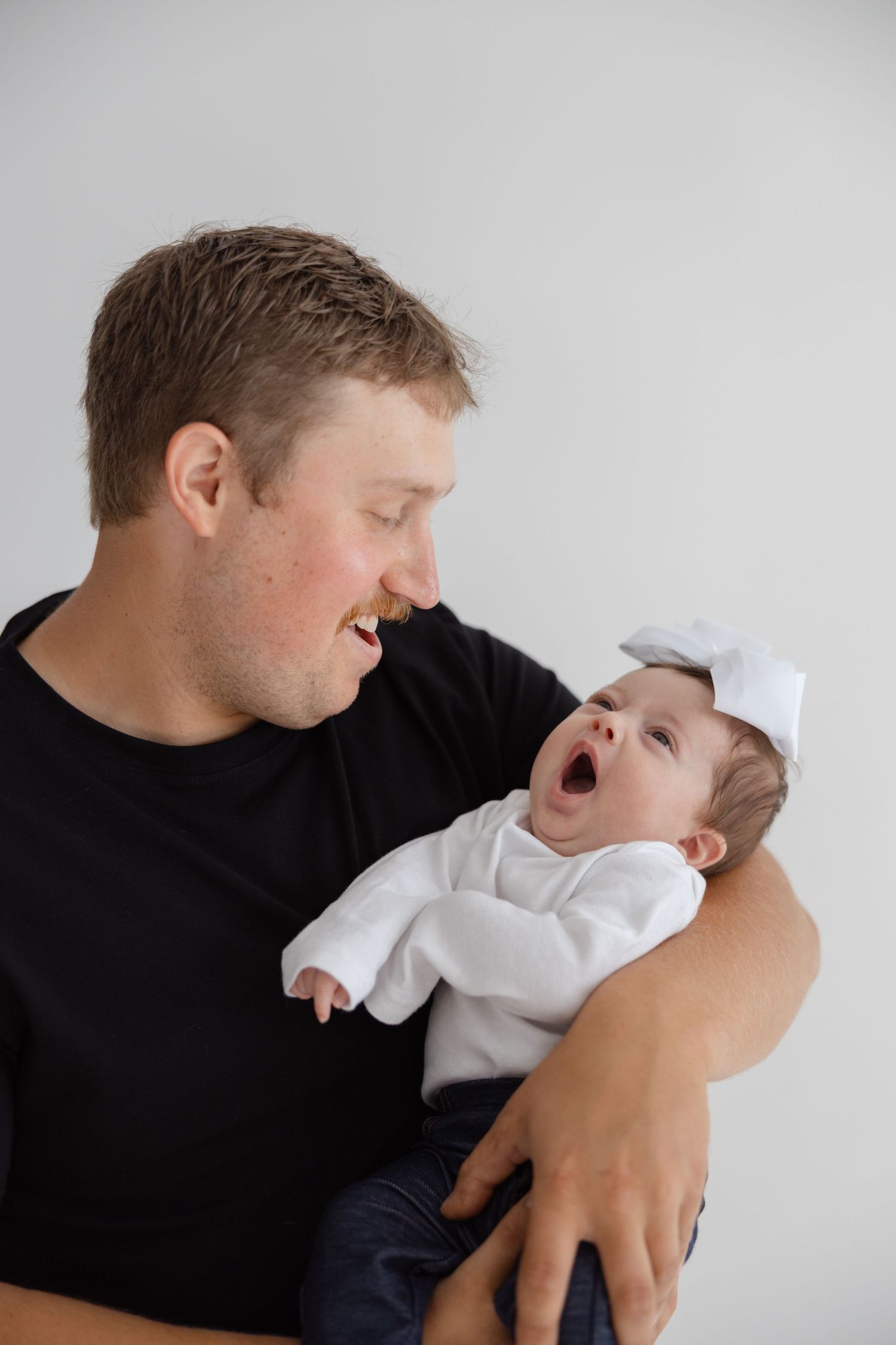 A person in a black shirt holds a laughing baby wearing a white outfit against a light gray background.