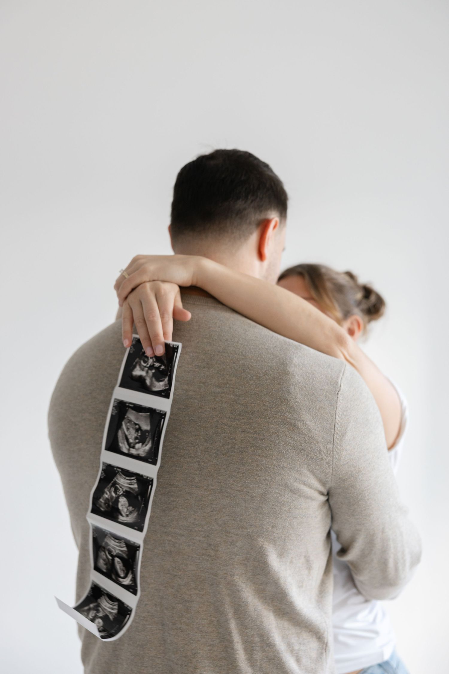 Couple embraces while holding ultrasound strip showing baby scans in emotional pregnancy announcement moment.