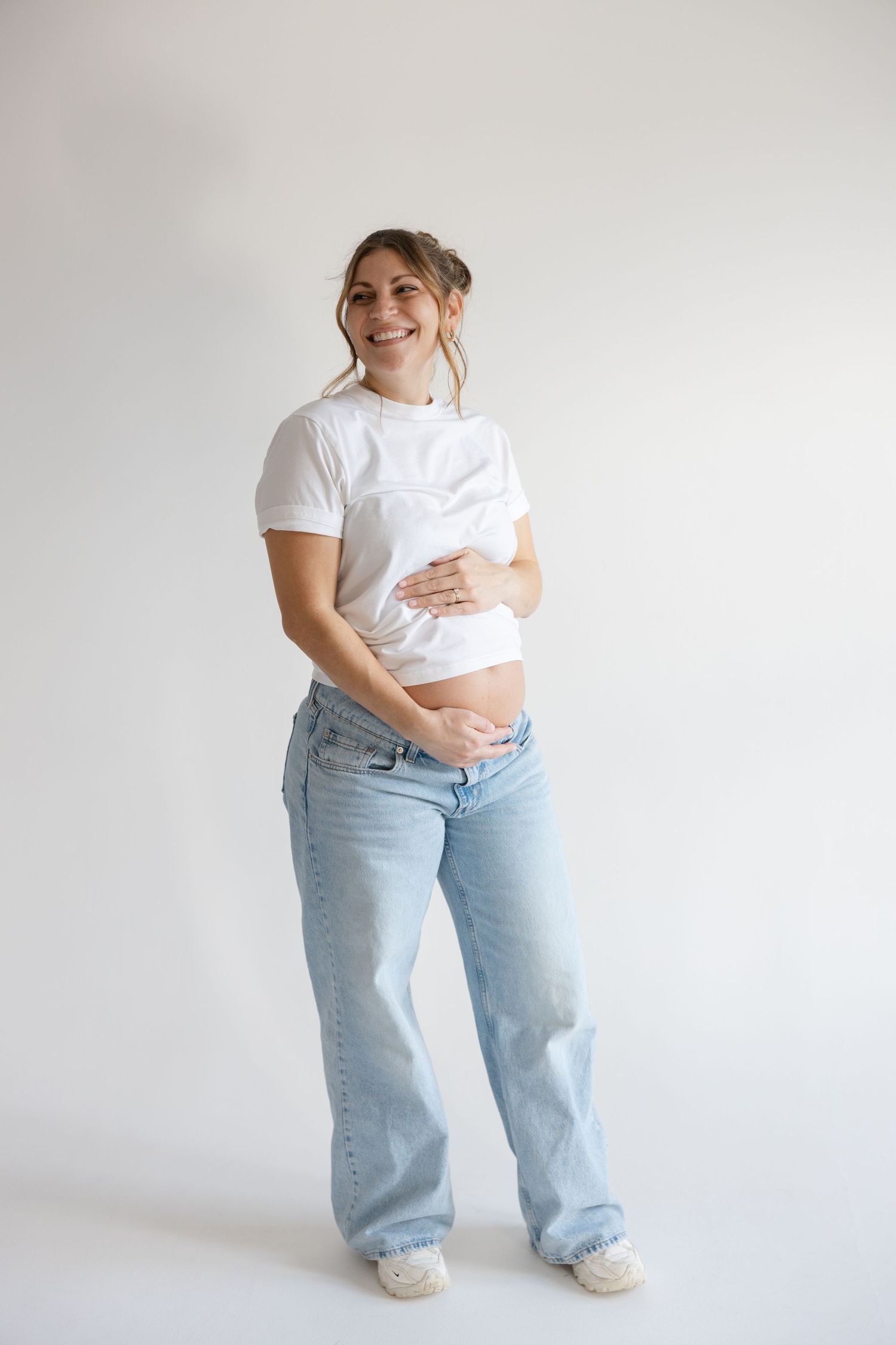 Expectant person in white t-shirt and light blue jeans poses against white background while holding baby bump.